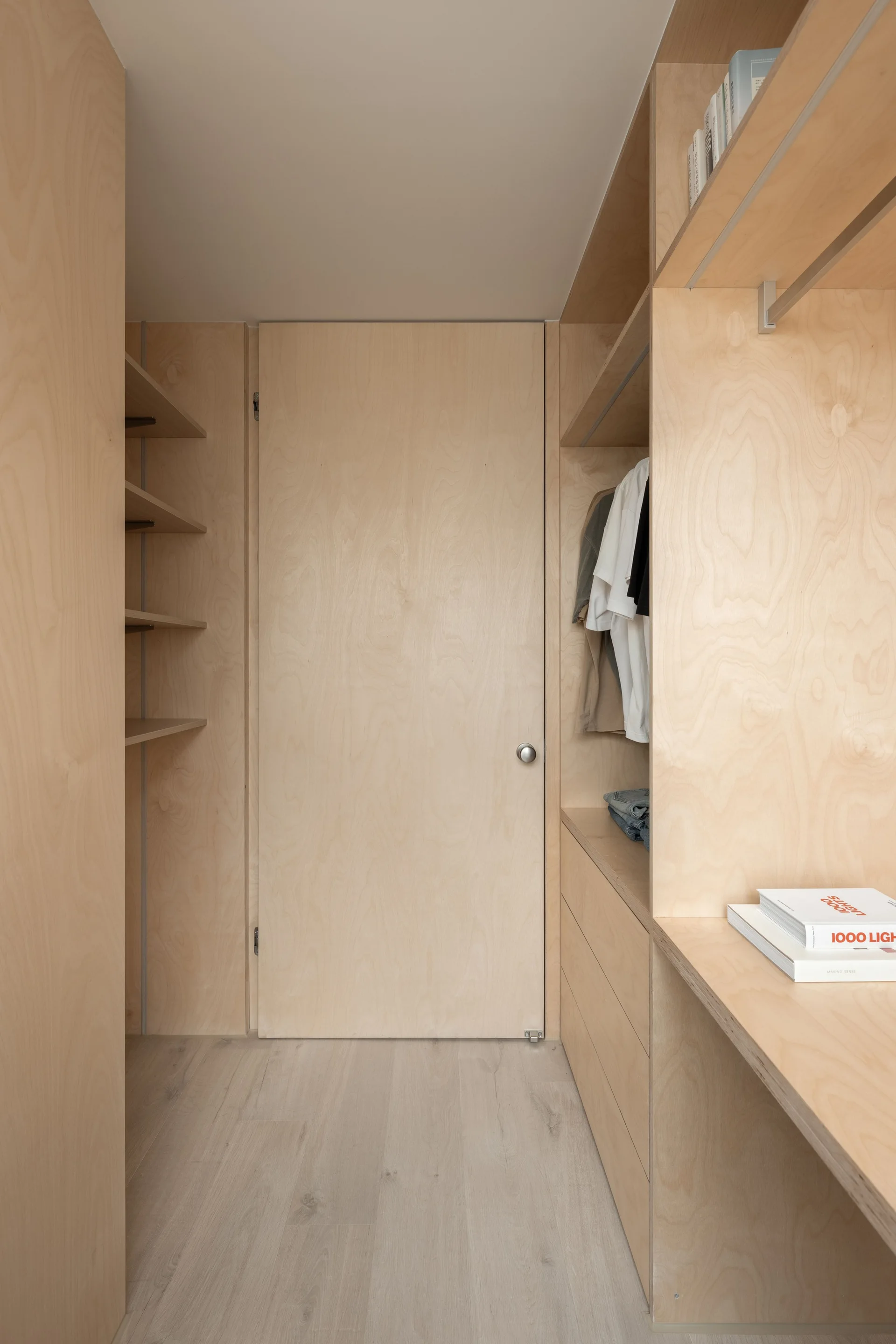 A minimalist walk-in closet features built-in plywood shelving, clothes hanging on a rod, and a matching wood-panel flush door.