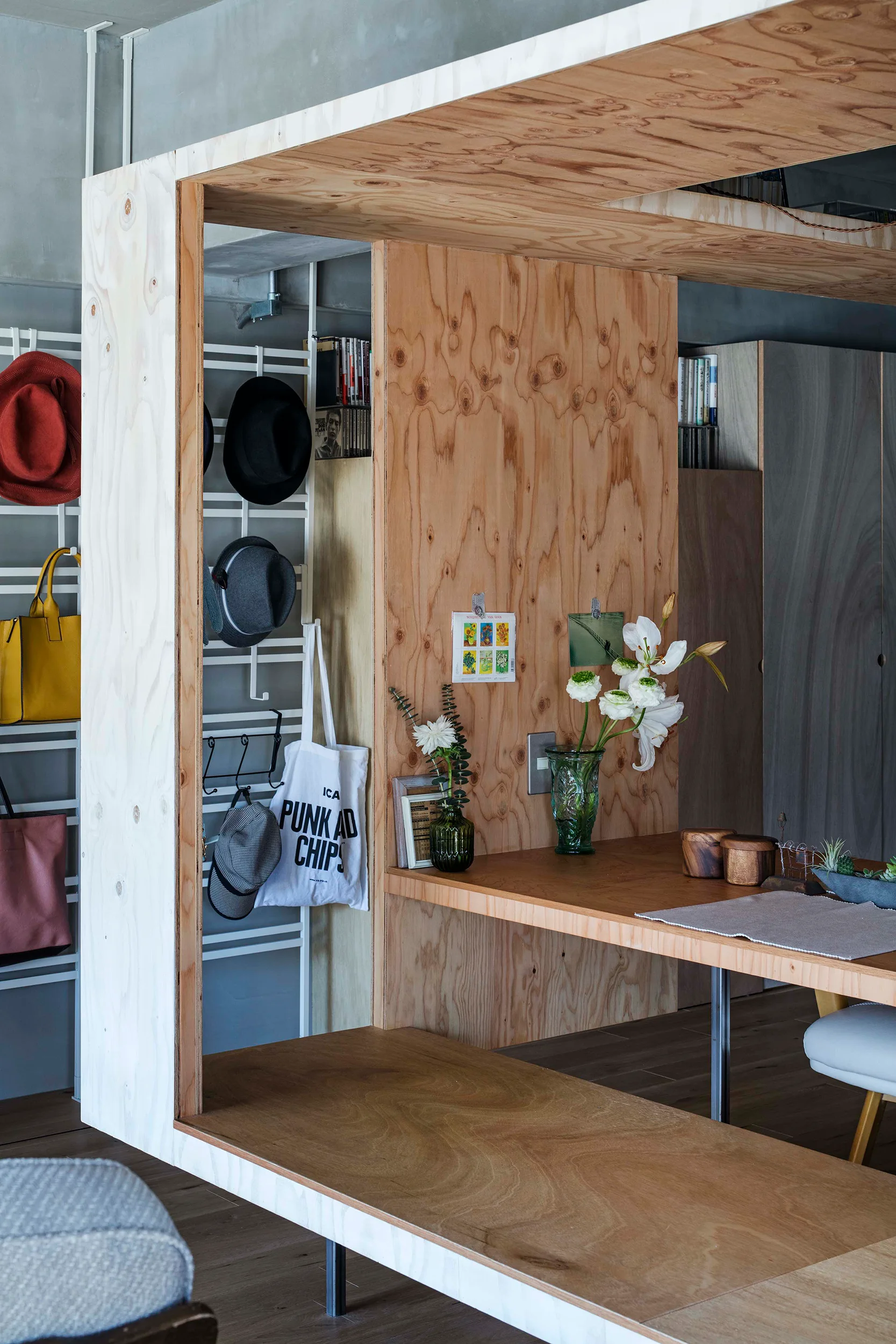 A minimalist interior features a custom plywood room divider with an integrated desk, alongside a metal rack holding hats and bags.
