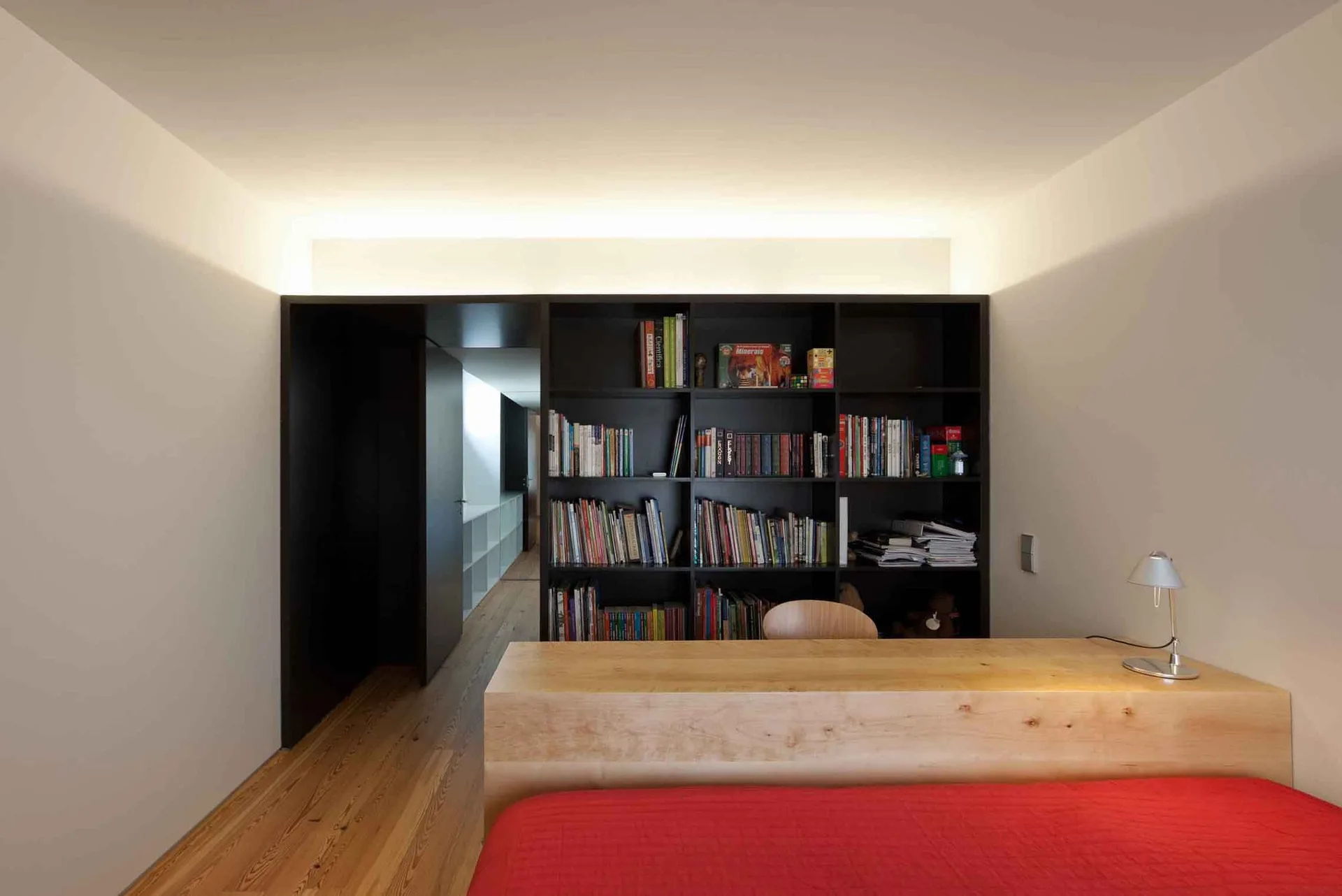 A minimalist bedroom features a red bed, a light wood desk that acts as a headboard, a dark built-in bookshelf, and warm ceiling cove lighting.