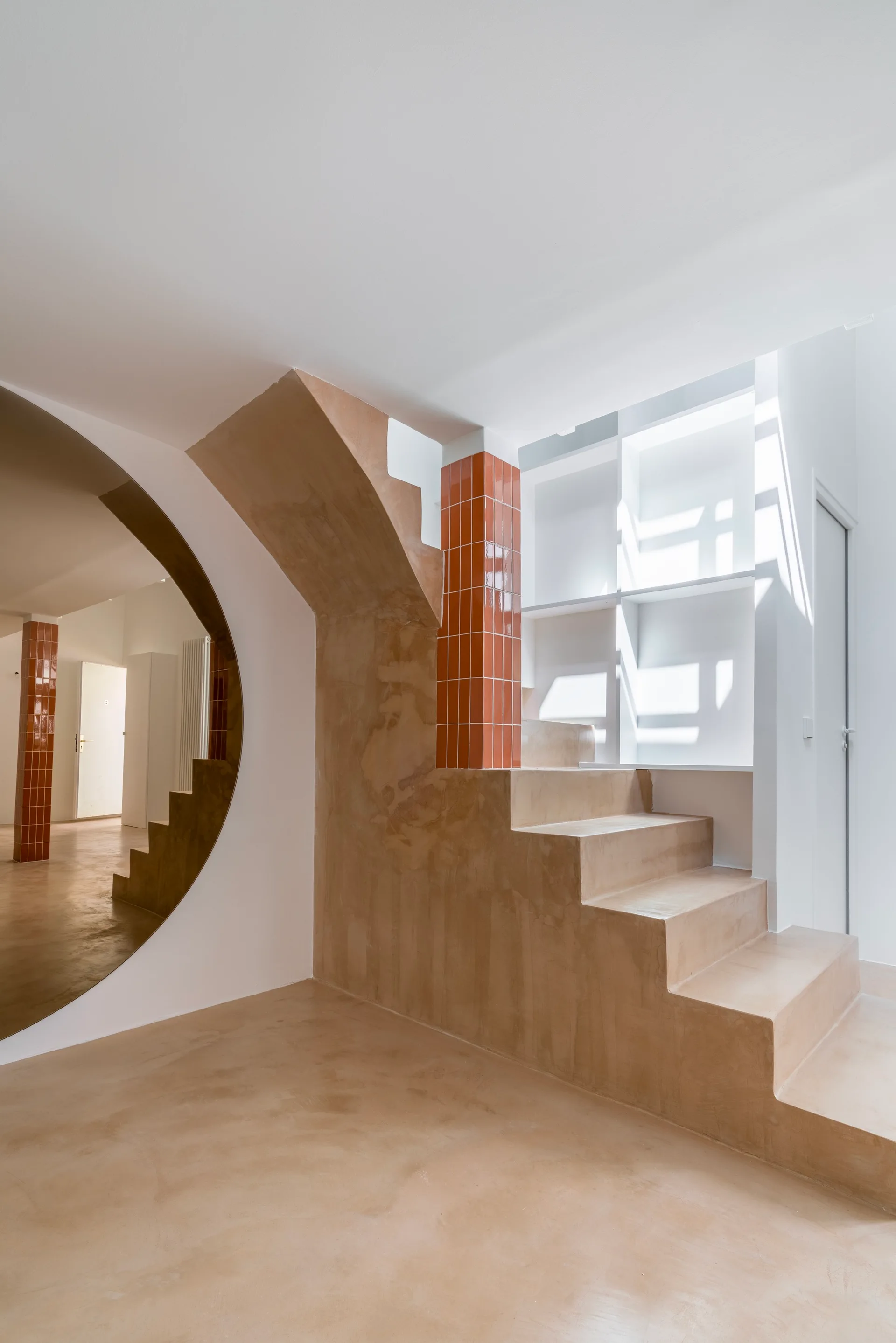 A minimalist beige plaster staircase stands next to a glossy orange tiled column and a large circular mirror reflecting a white hallway.
