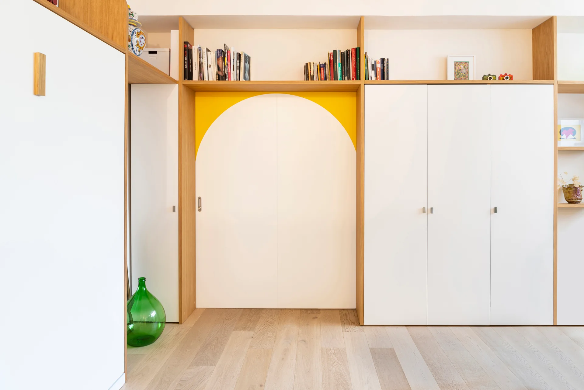 Floor to ceiling built-in white cabinets featuring light wood shelving, a central painted yellow arch design, and matte light wood flooring.