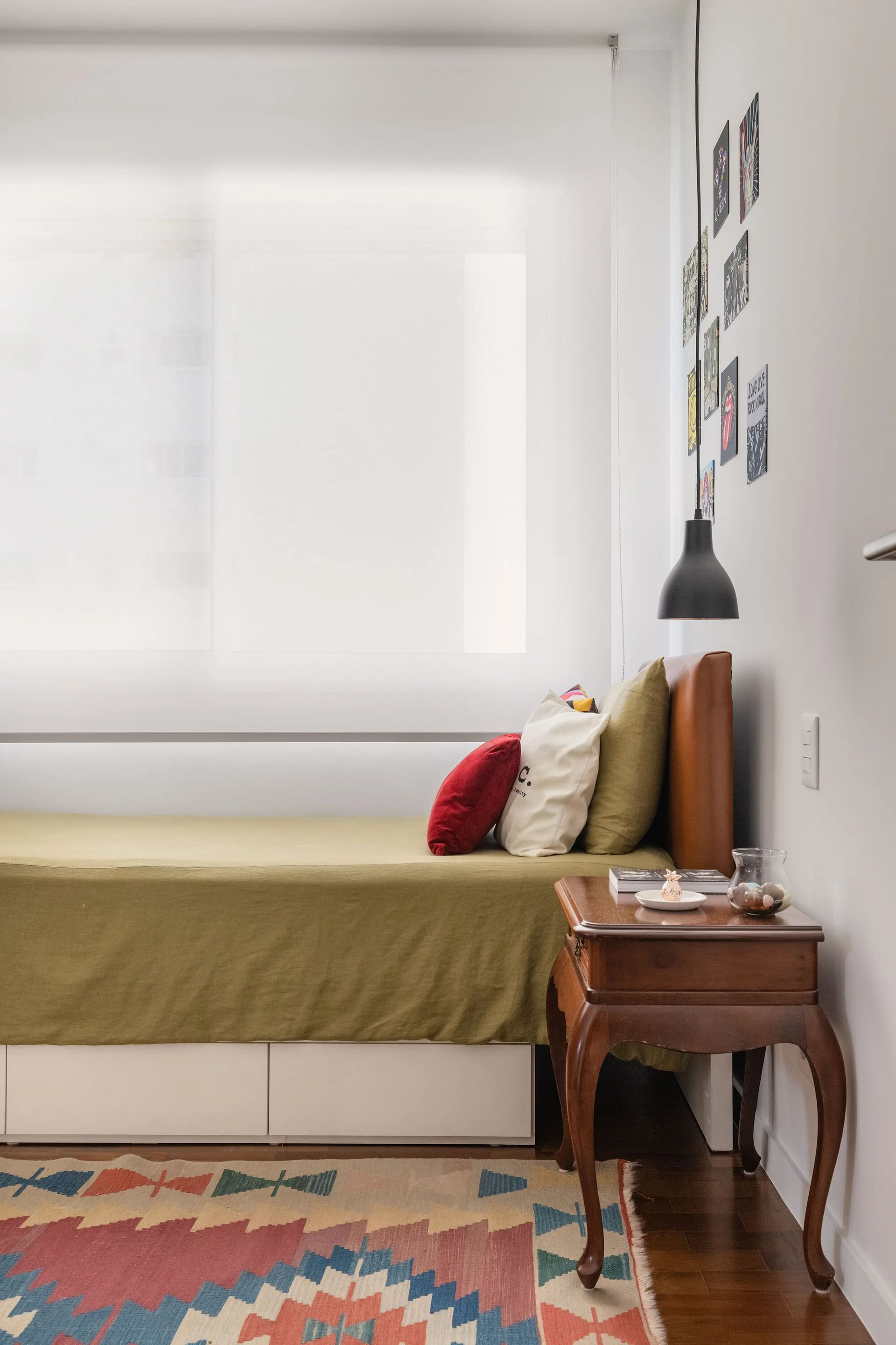 A compact bedroom corner featuring an olive green bed, a wooden side table with cabriole legs, a black pendant light, and a colorful geometric rug.