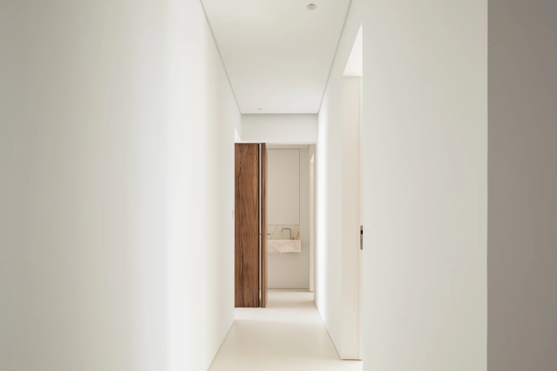 A minimalist white hallway leads to a bathroom area with a tall wood door and a wall-mounted stone washbasin.