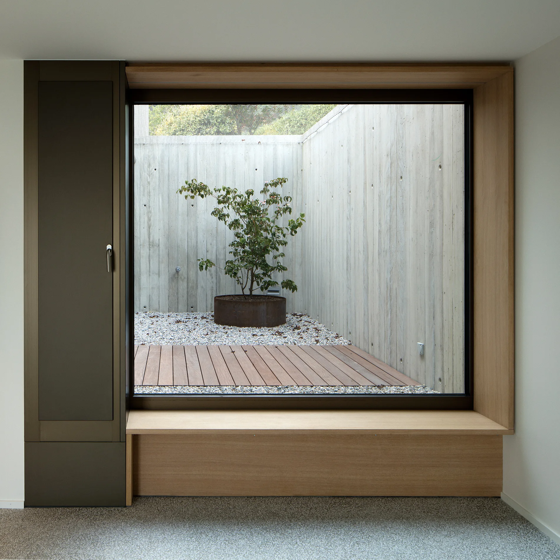 A large square window with a light wood frame and built-in bench looks out to an enclosed concrete courtyard with a potted tree and gravel.