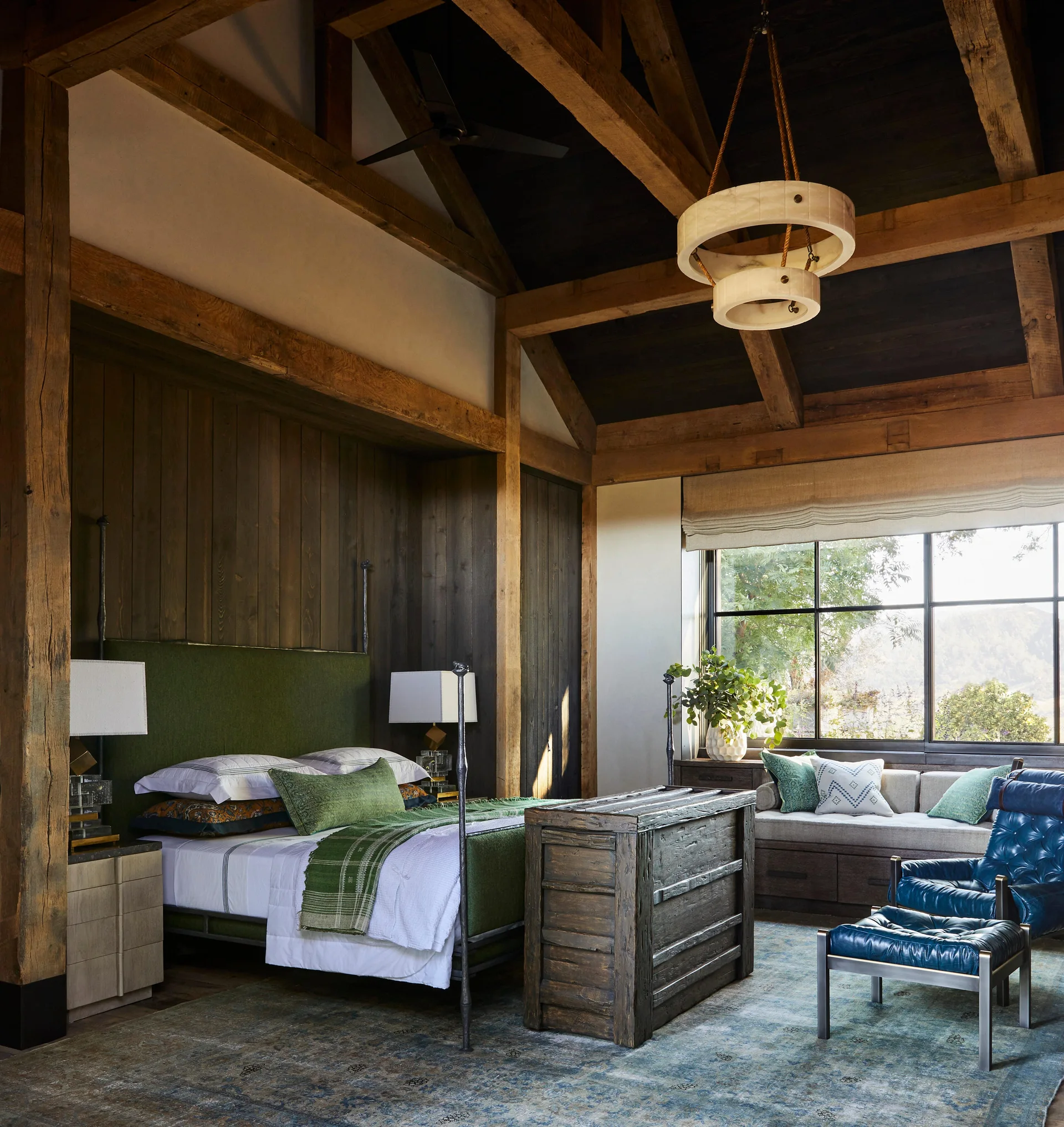 A rustic bedroom features exposed timber beams on a vaulted ceiling, a bed with a green headboard, a wooden chest, and a blue leather armchair.