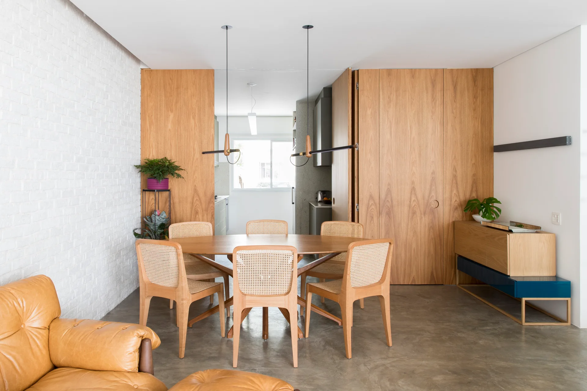 A dining room features a wooden table with six cane-back chairs set against a white brick wall and warm wood paneling.