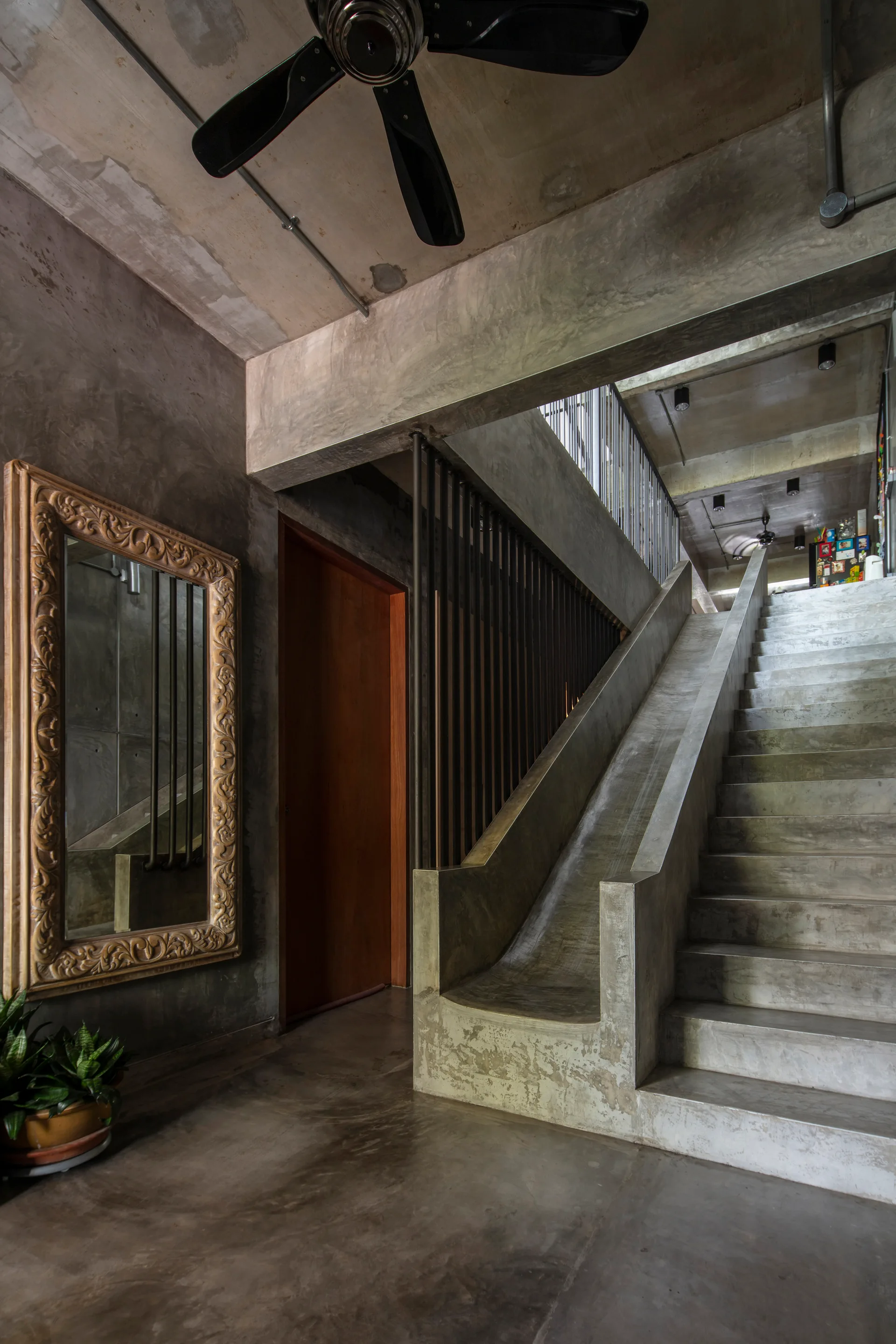 An industrial hallway features a raw concrete staircase with a built-in concrete slide, a warm wood door, and an ornate carved mirror on the wall.