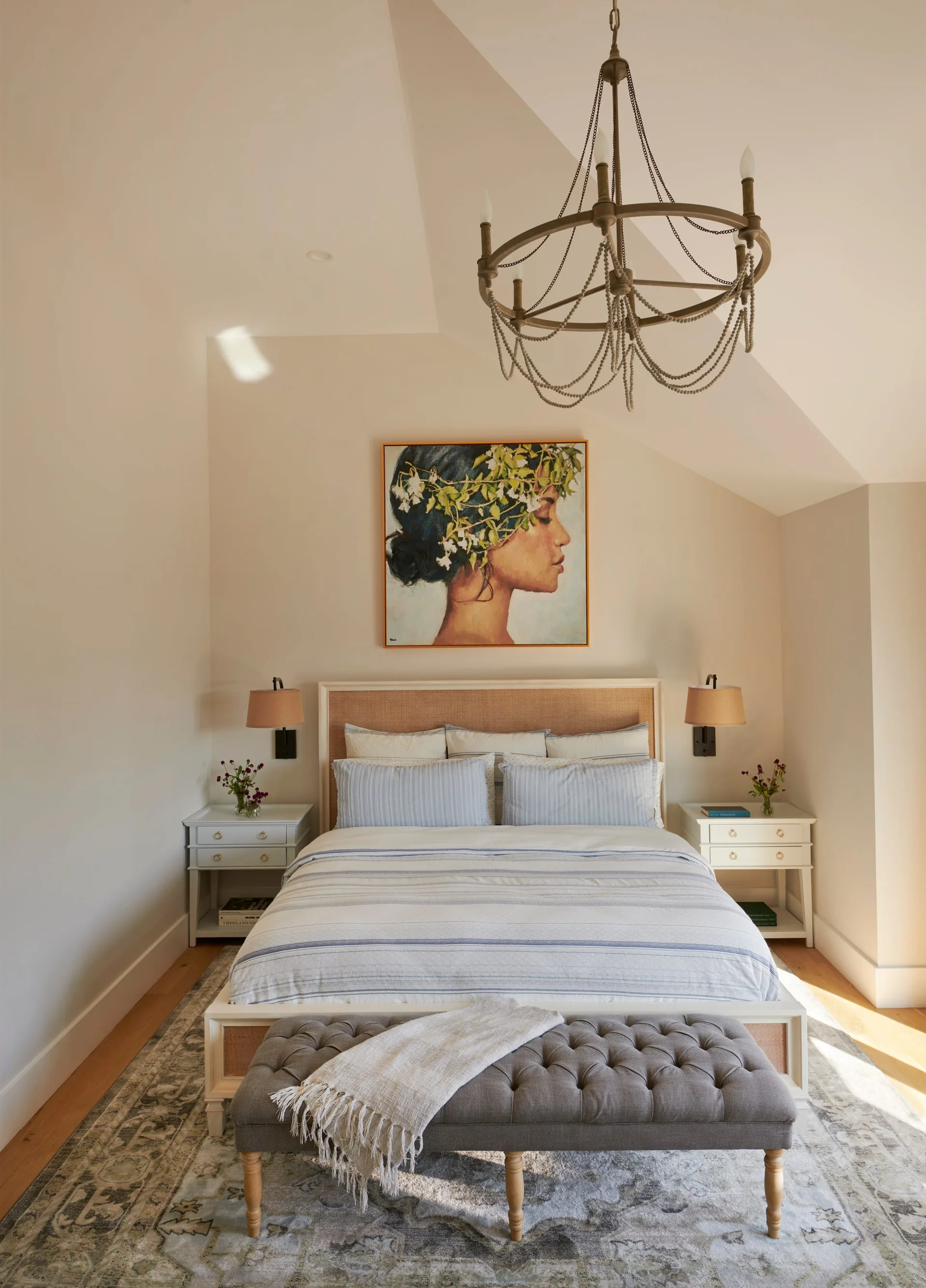 A bedroom features a bed with striped bedding, a gray tufted bench, white nightstands, and a beaded chandelier under a sloped ceiling.