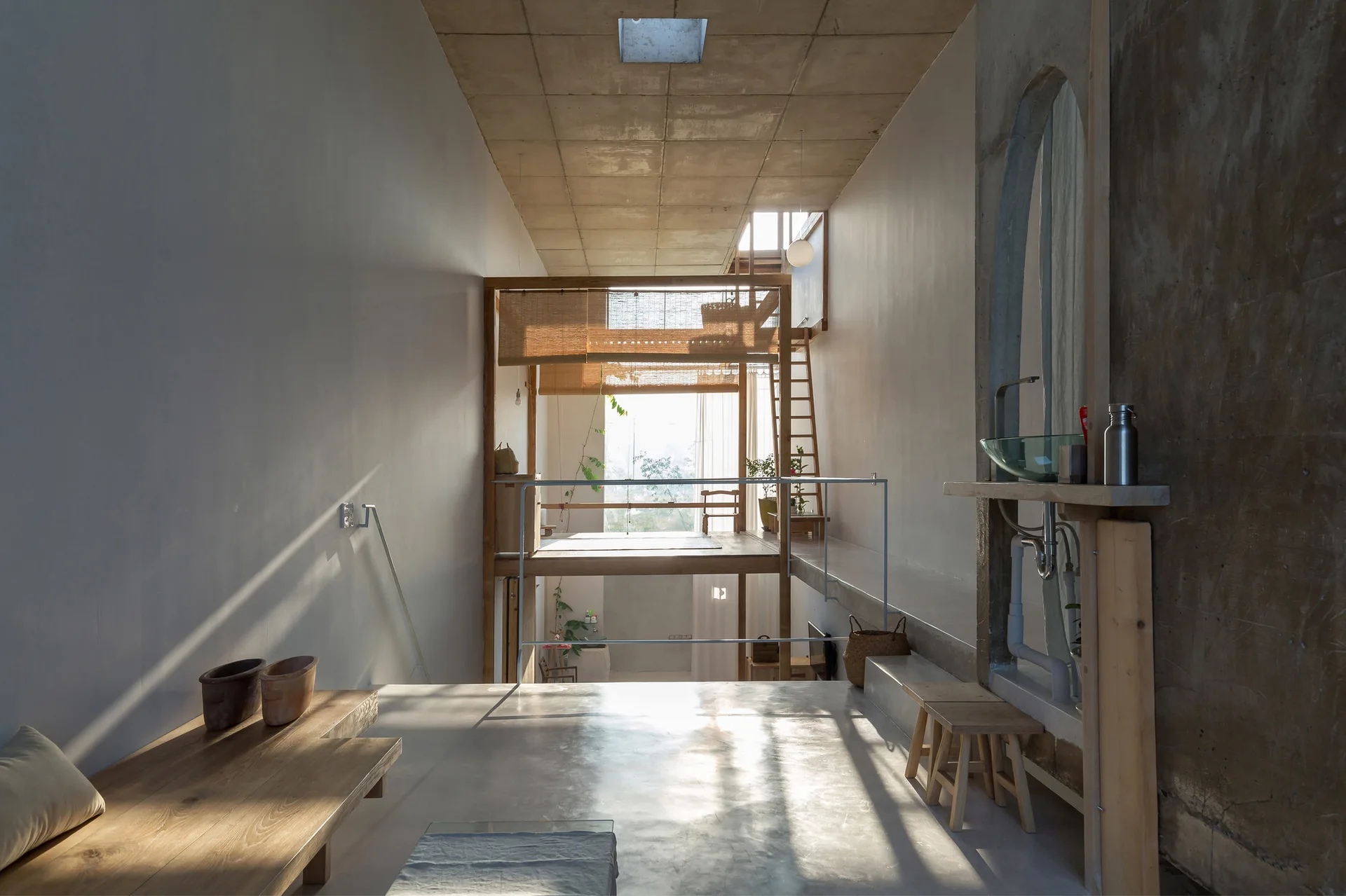 A minimalist loft features polished concrete floors, a concrete grid ceiling, a raised wooden platform with woven blinds, and a long wooden bench.