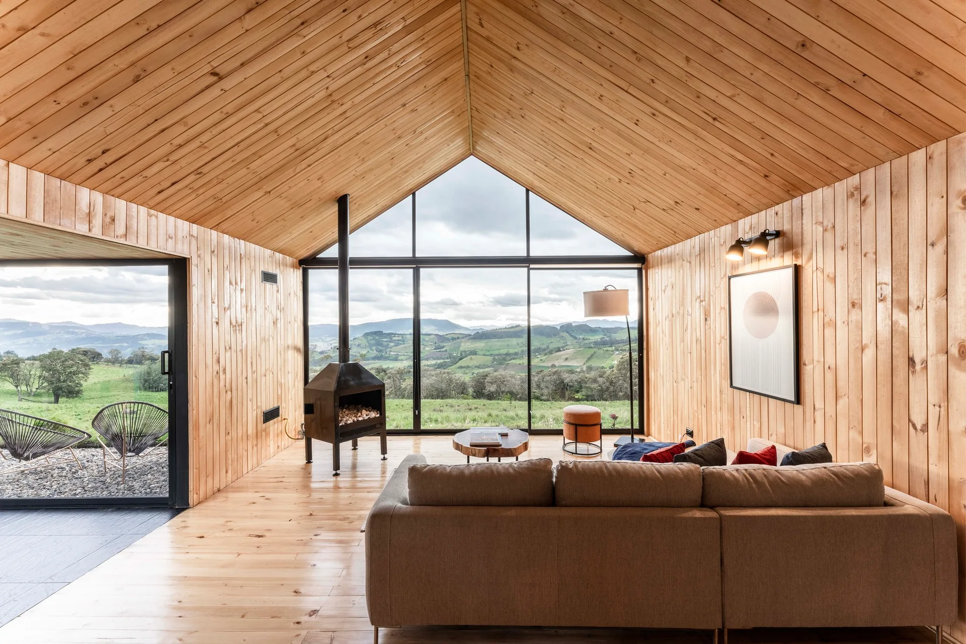 A modern cabin living room features light wood paneled walls, a vaulted ceiling, a tan sofa, a black fireplace, and a large floor-to-ceiling window.