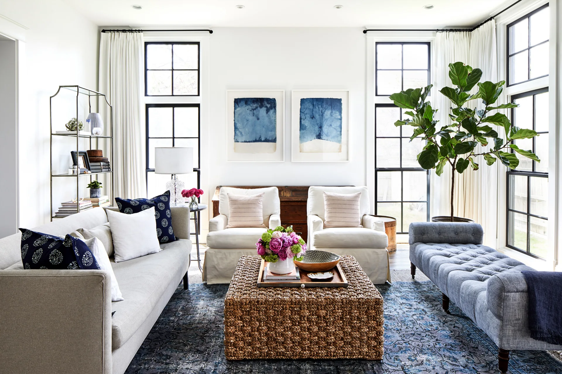 A bright living room with a grey sofa, two white slipcovered armchairs, and a large woven coffee table on a patterned blue rug.
