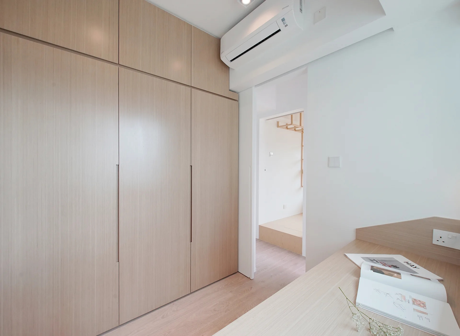 A minimalist bedroom features a large floor-to-ceiling light wood closet next to a doorway and a built-in study desk with an open book.
