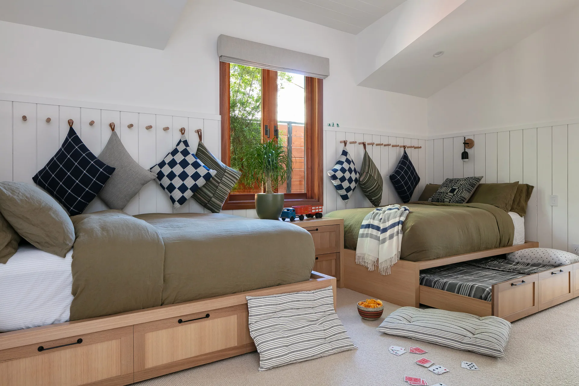 A shared children's bedroom featuring two light wood trundle beds, olive green duvets, and patterned pillows hanging from wooden wall peg hooks.