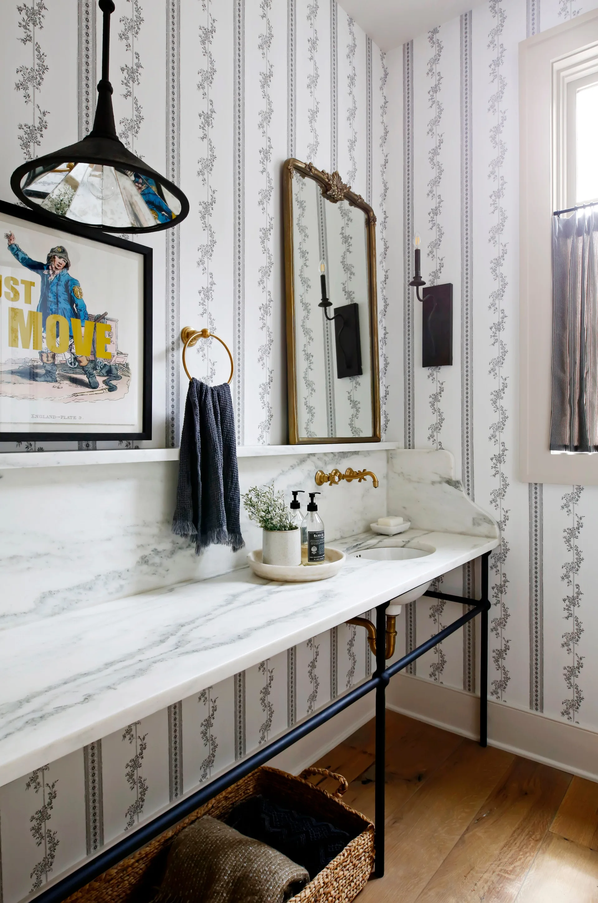 A powder room vanity features a marble console countertop, a gilded mirror, brass fixtures, and black-and-white striped floral wallpaper.