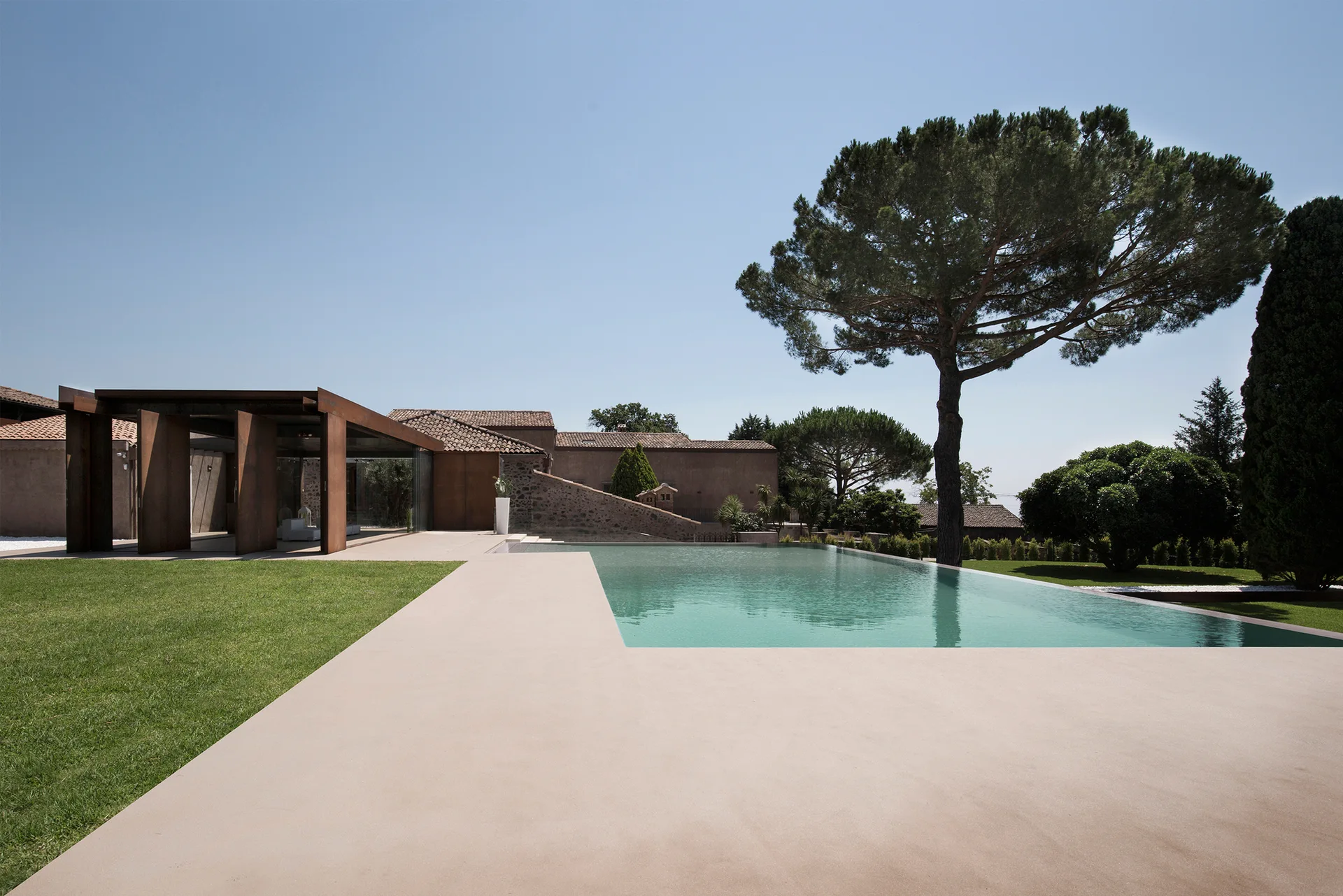 A rectangular swimming pool with clear turquoise water next to a smooth concrete deck, a weathered metal pergola, and green lawns.