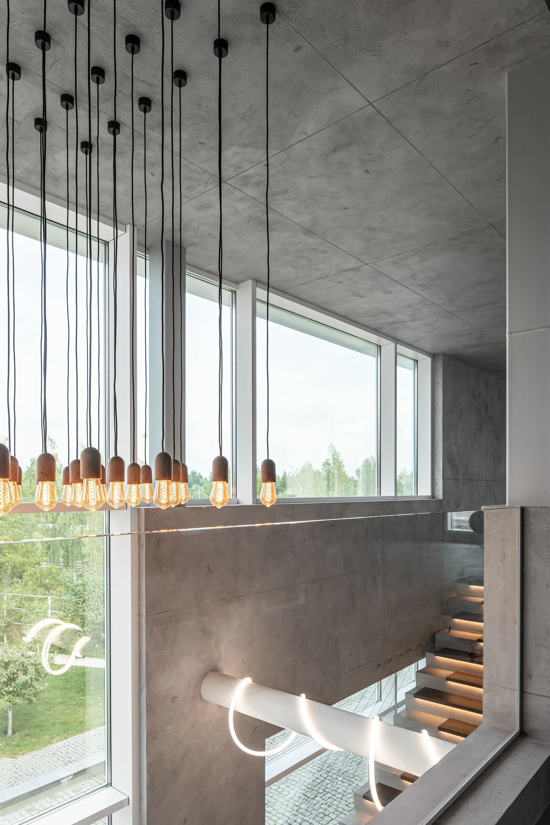 A modern concrete stairwell features a massive cluster of hanging Edison bulb pendant lights in front of a large window.