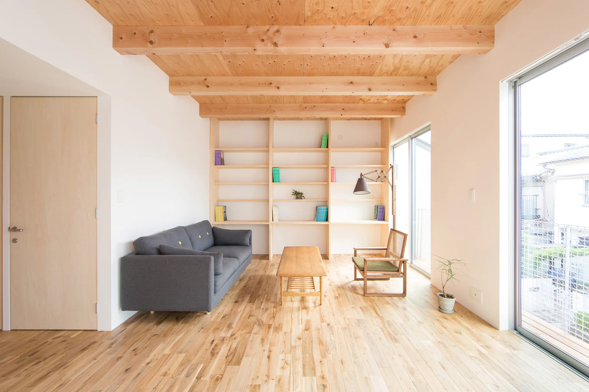 A minimalist living room features a grey sofa, a wooden coffee table, and large built-in shelving beneath an exposed timber ceiling.