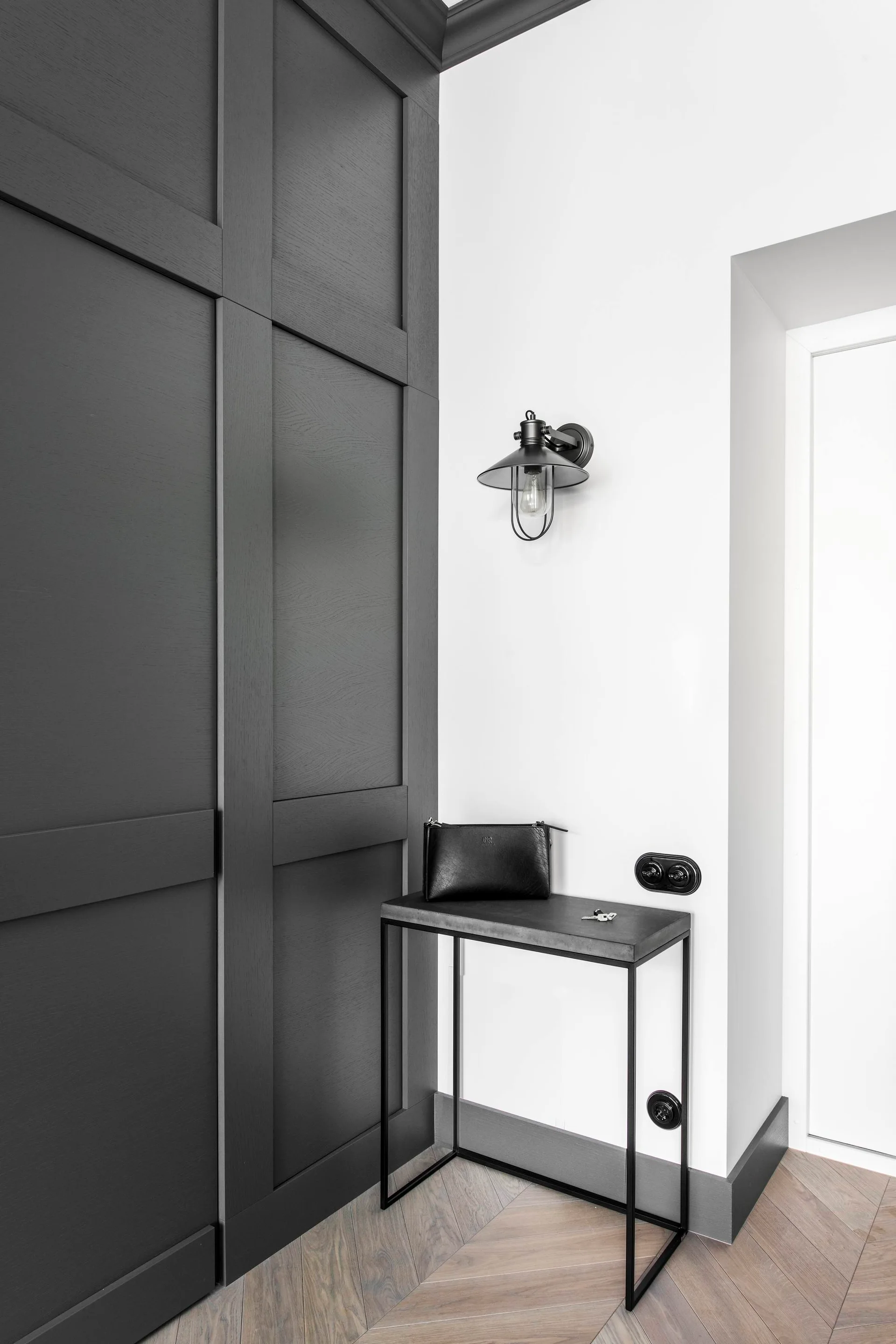 A minimalist entryway features dark gray paneled built-in cabinets, a slim black metal console table, and a wall-mounted industrial sconce.