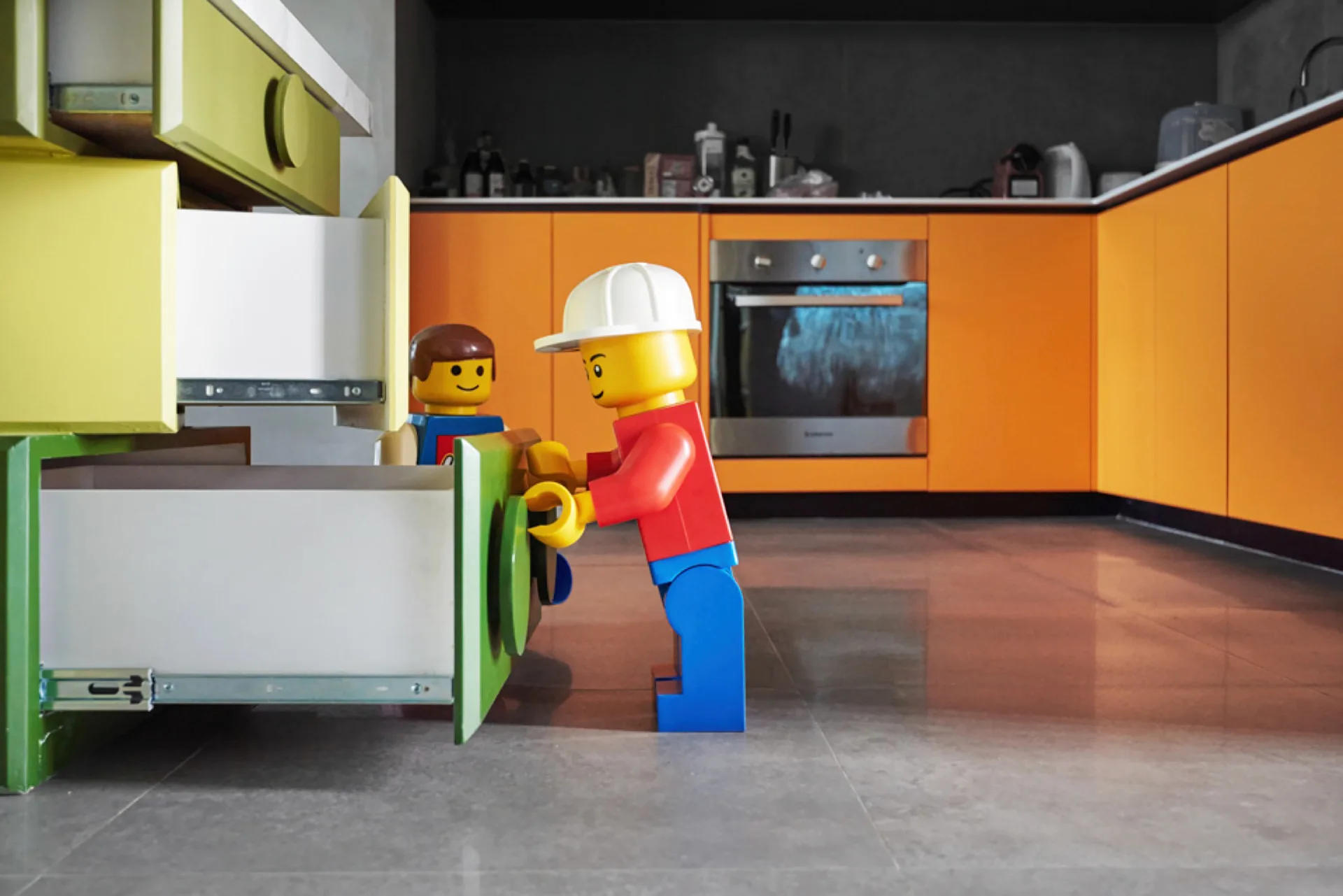 A modern residential kitchen with matte orange cabinets, a built-in steel oven, gray floor tiles, and colorful drawers in the foreground.