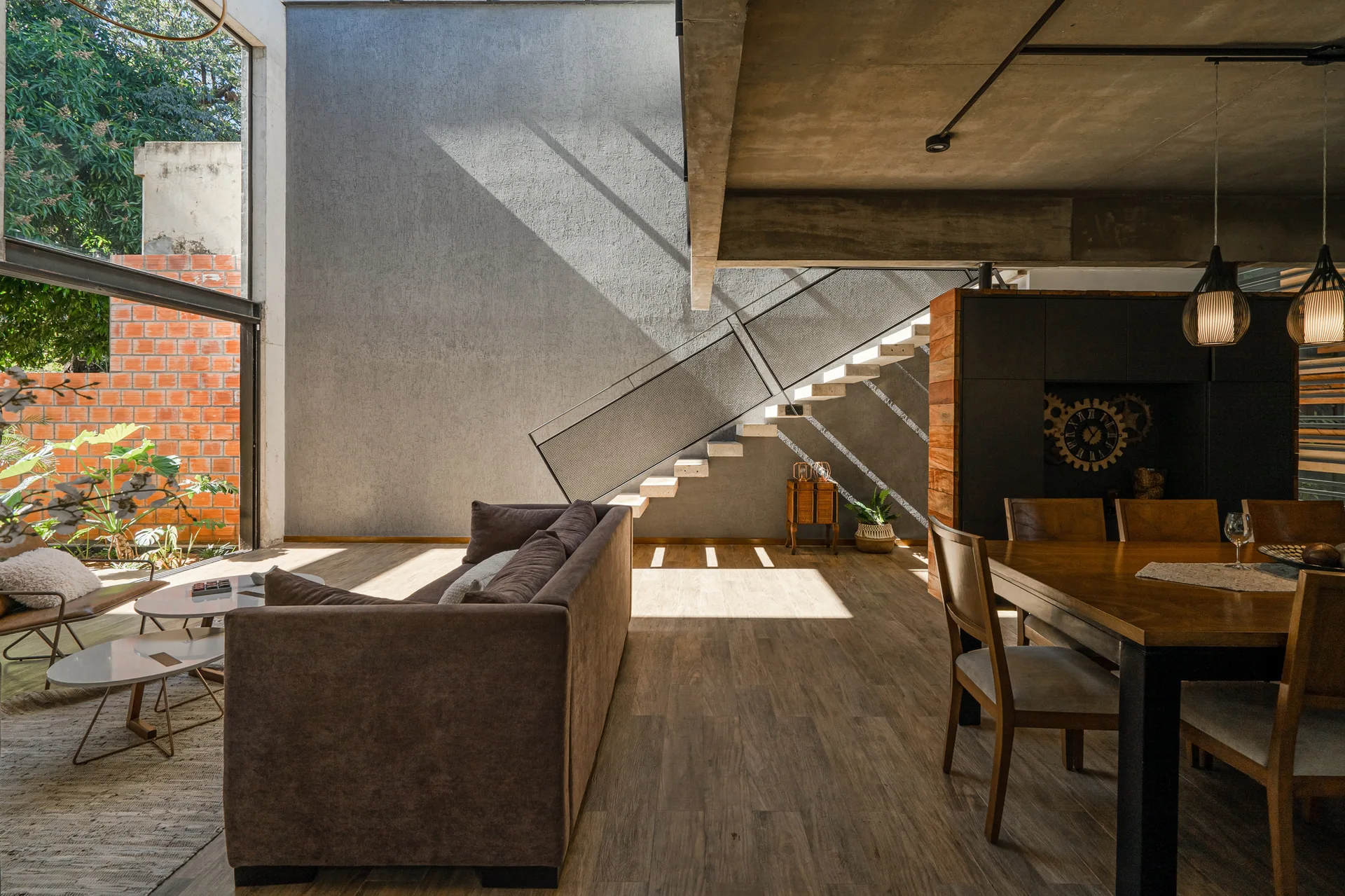 An open plan living area features a dark brown fabric sofa, a wood dining table, and concrete floating stairs against a textured grey plaster wall.