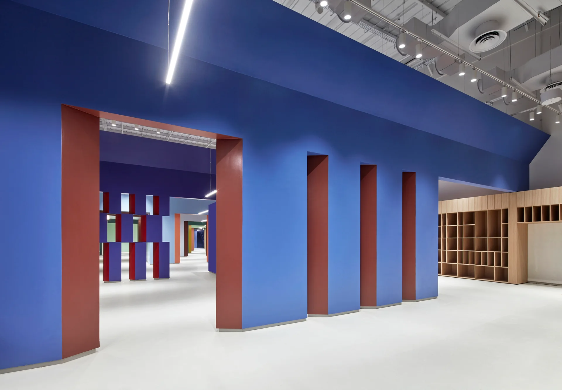 A spacious open-plan interior featuring bold blue architectural walls with deep red openings alongside a light wood grid shelving unit.