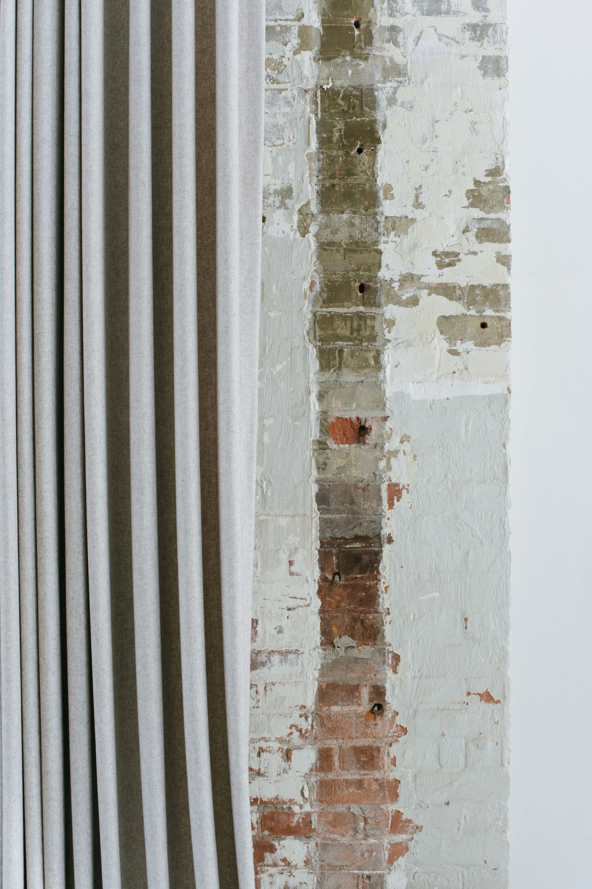 A pleated light gray fabric curtain hangs next to a rustic, partially painted brick wall with exposed red and beige brickwork.
