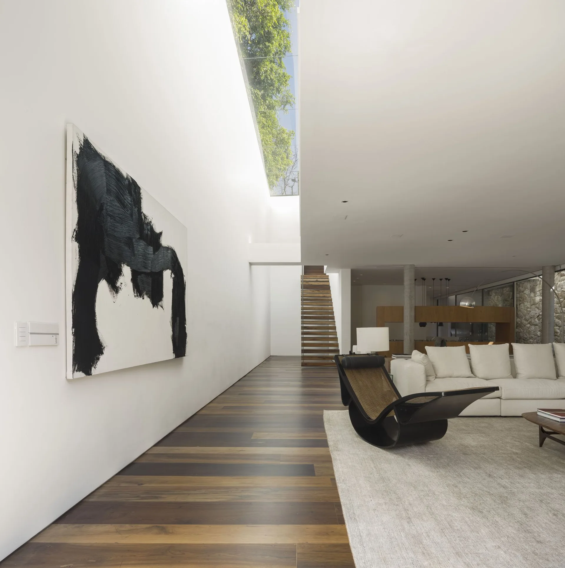 A minimalist living room features a long skylight, a large black and white abstract painting, a curved lounge chair, and a white sofa.