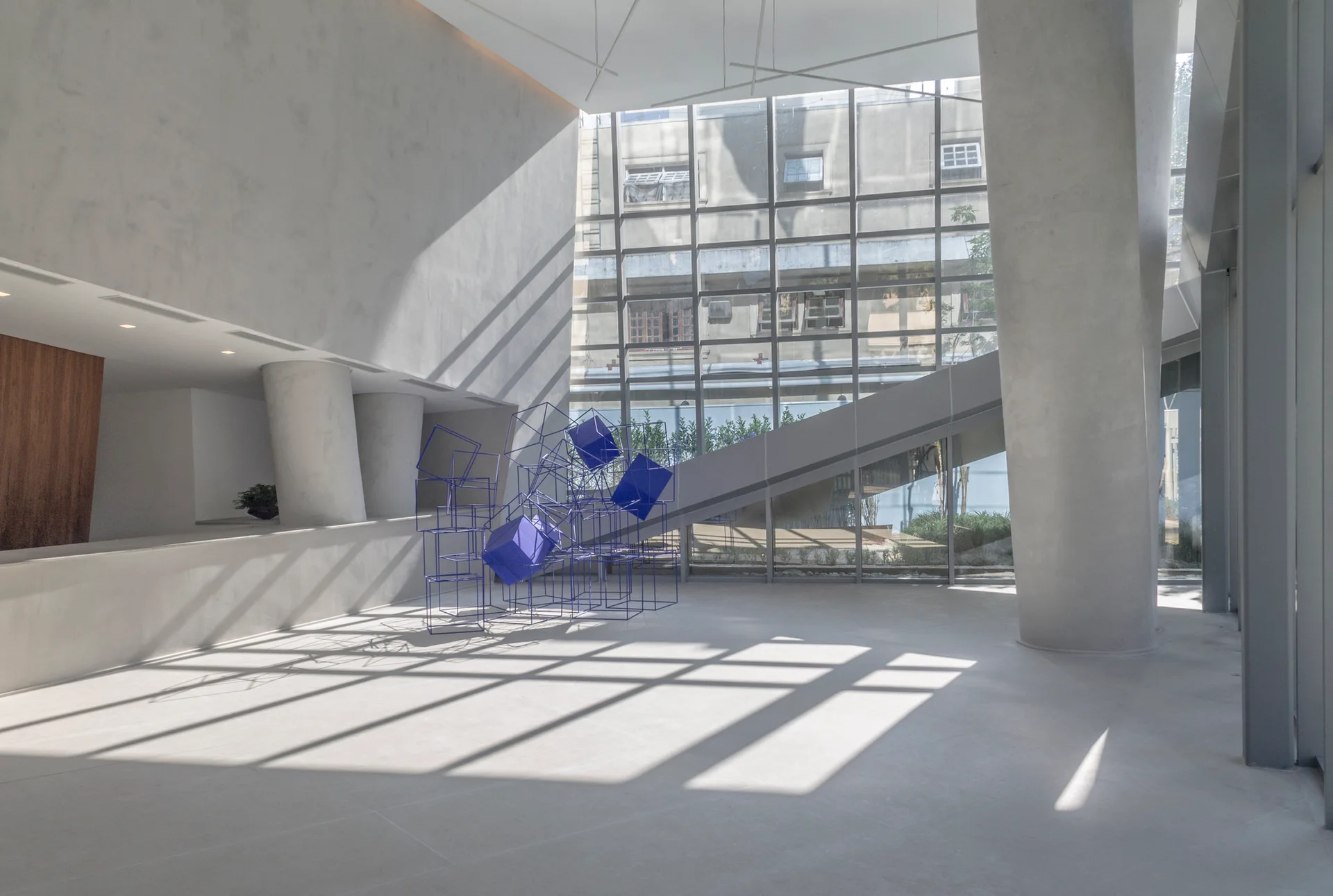 A spacious minimalist lobby features smooth concrete columns, a glass facade, and a blue geometric wireframe sculpture on a light grey floor.