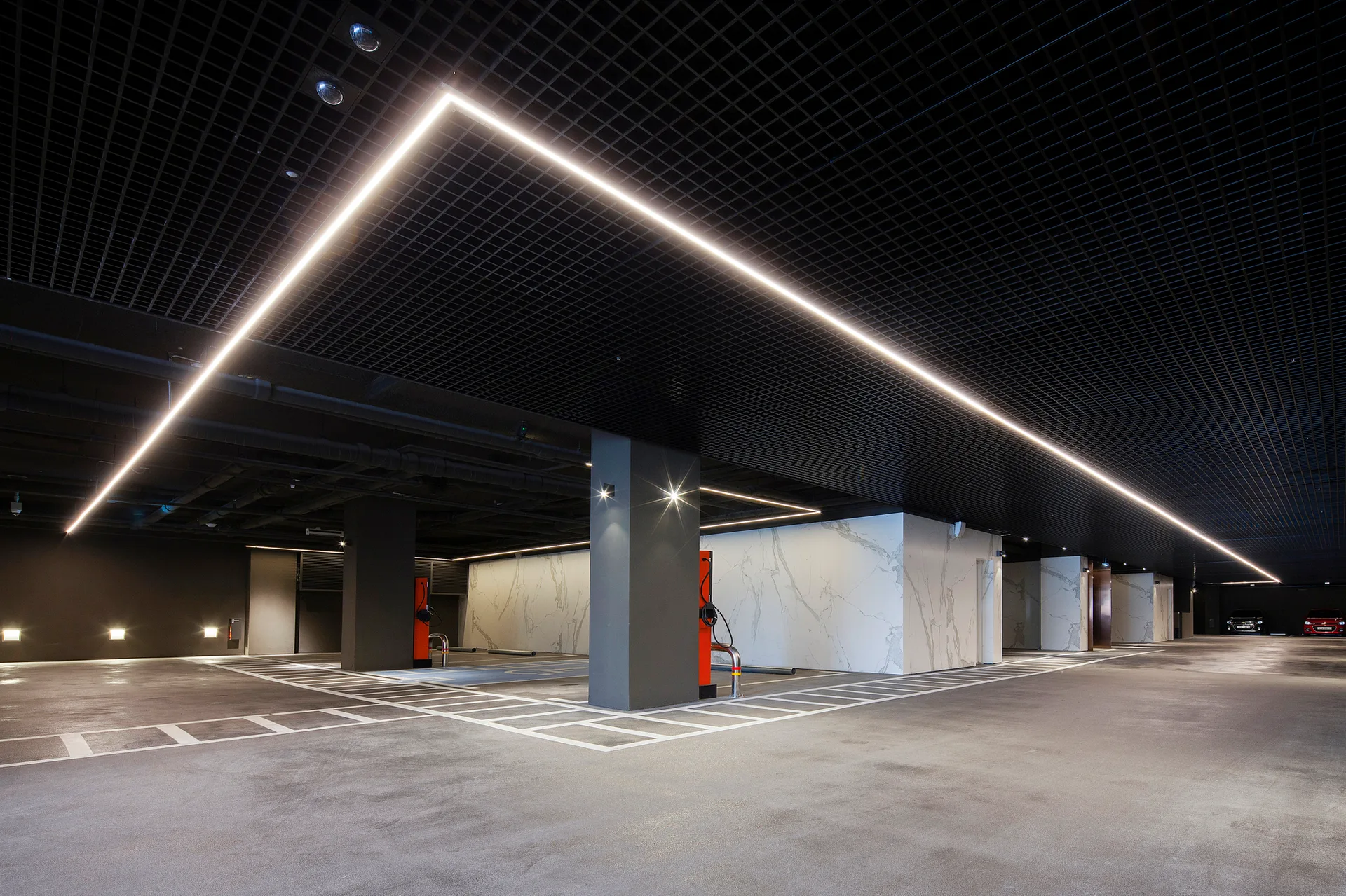 An underground parking garage featuring a black grid ceiling, linear LED strip lights, concrete floors, and veined white marble wall cladding.