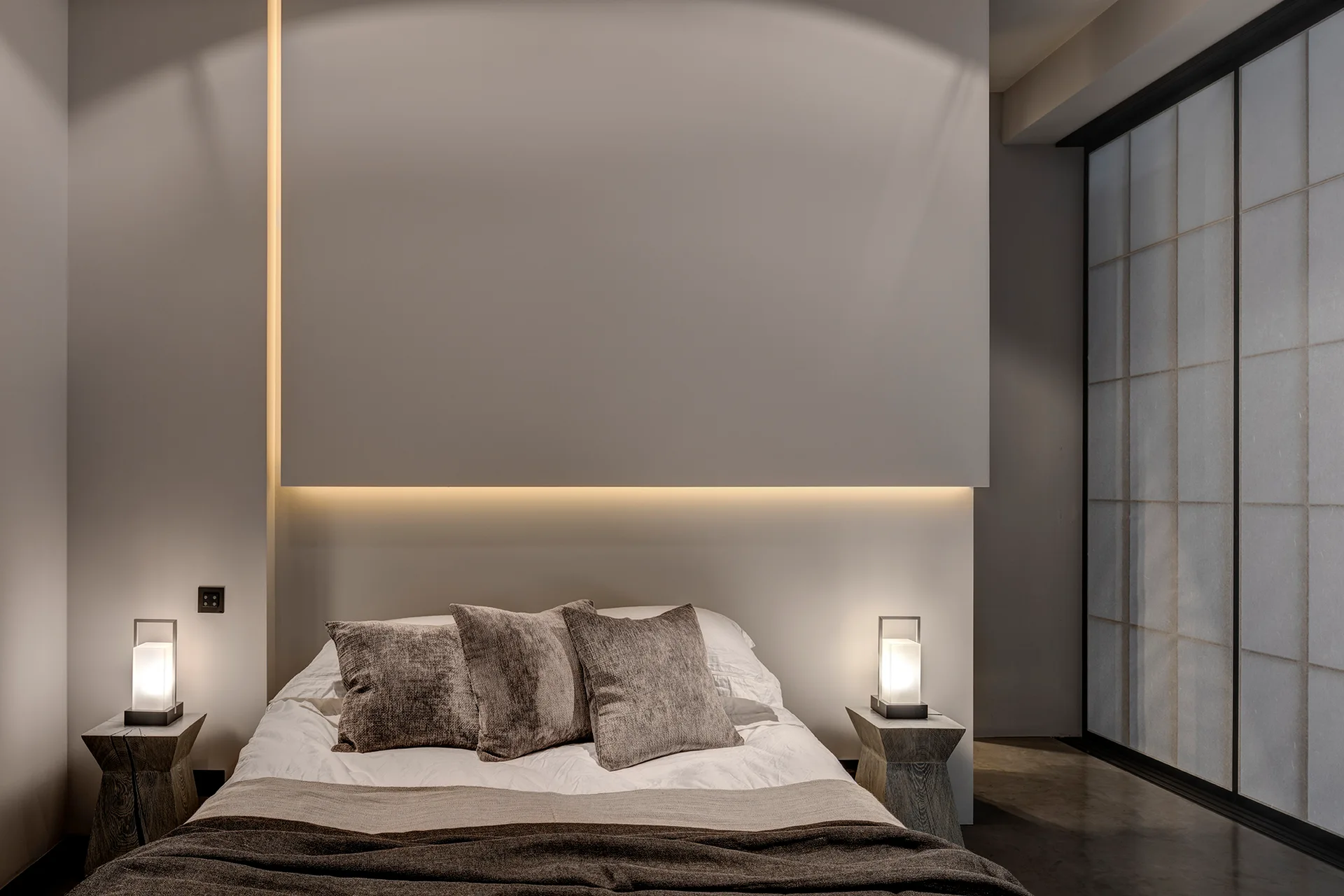 A minimalist bedroom features a bed centered under a backlit wall panel, flanked by geometric wooden side tables with glowing lantern-style lamps.
