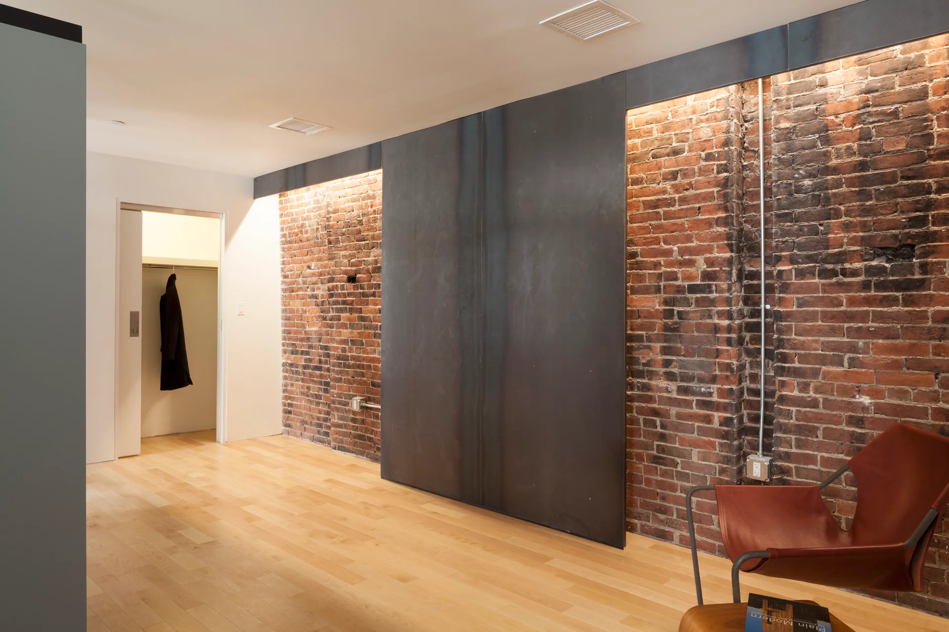 An industrial hallway features an exposed red brick wall illuminated by warm cove lighting next to a large dark metal sliding door and light wood flooring.