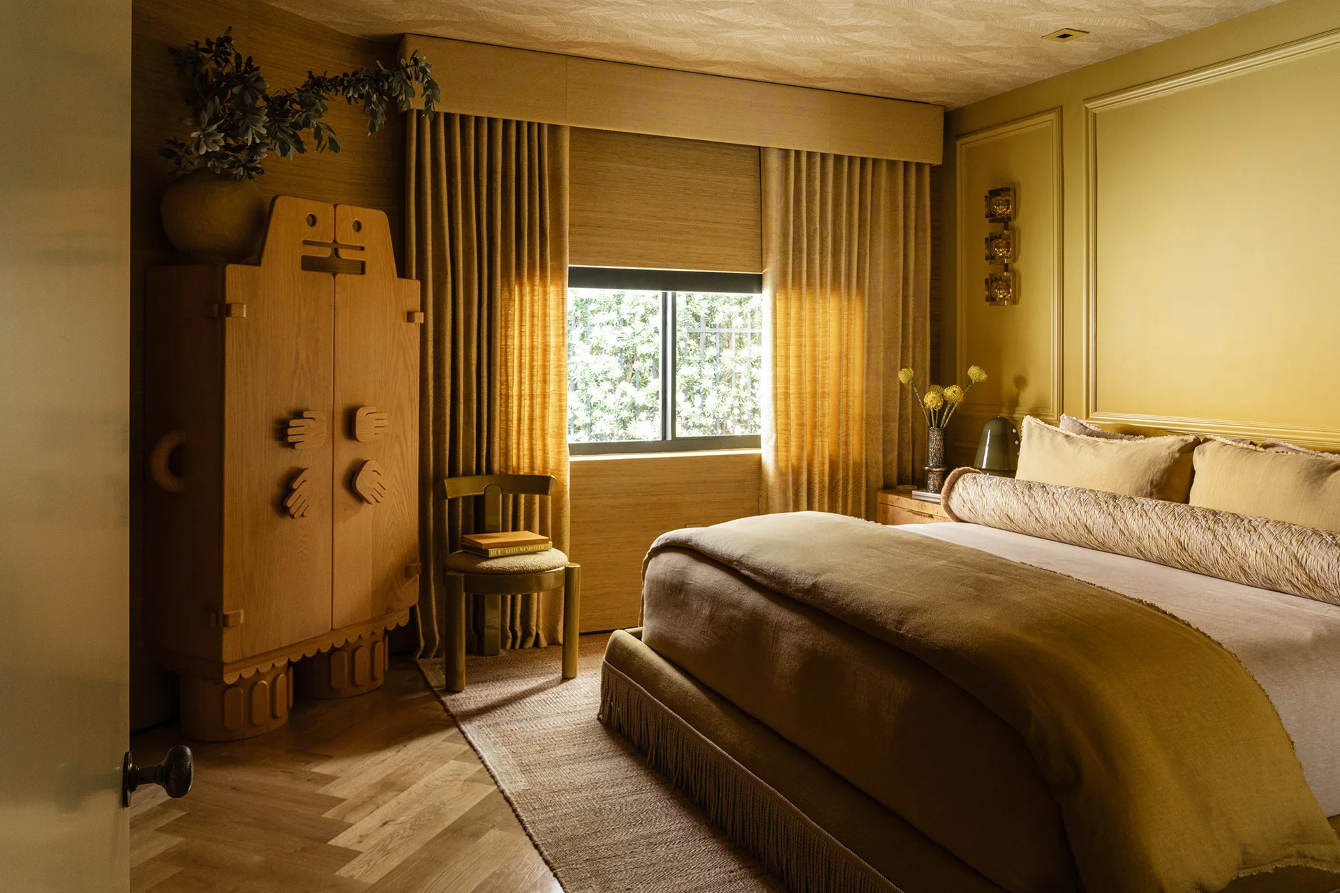 A warm ochre bedroom features an upholstered bed with fringe trim, a custom sculptural wooden cabinet, and floor-length matching curtains.