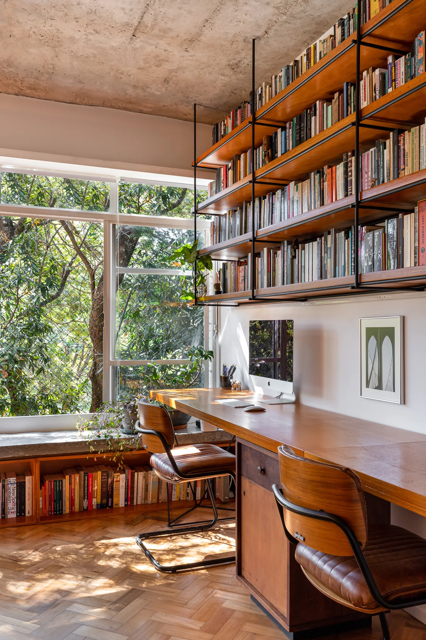 A home office features a long wooden double desk, vintage chairs, and a large wall-mounted bookshelf next to a window showing green trees.