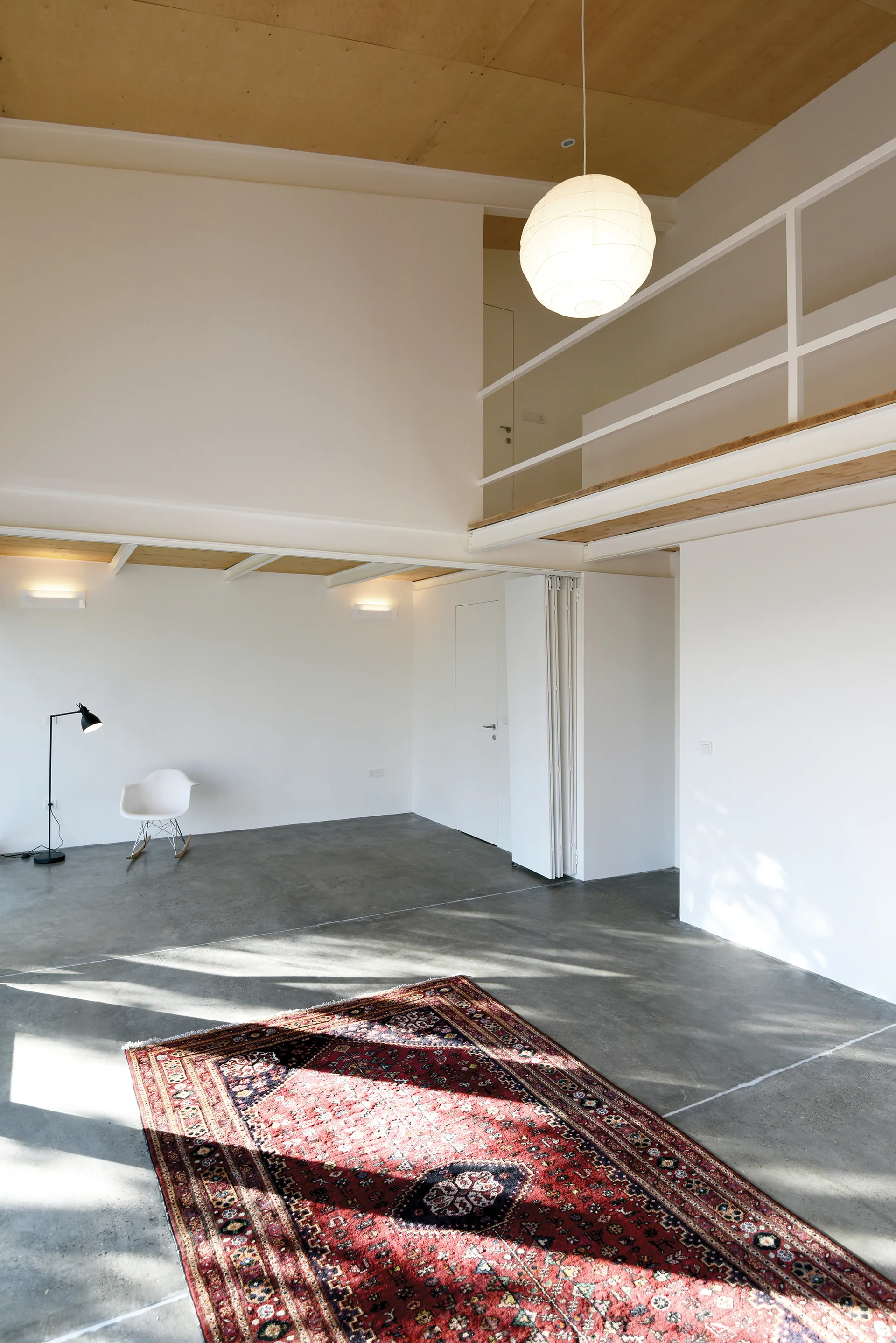 A spacious minimalist loft features a polished concrete floor, a white mezzanine level, and a large red patterned area rug.