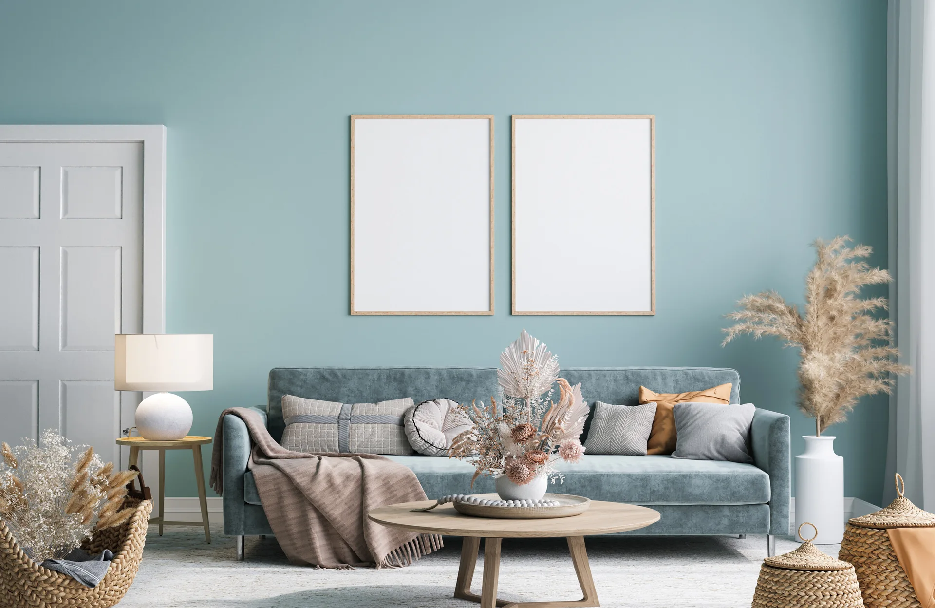 A light blue velvet sofa sits against a matching blue wall under two empty wooden frames, flanked by a round coffee table and wicker baskets.