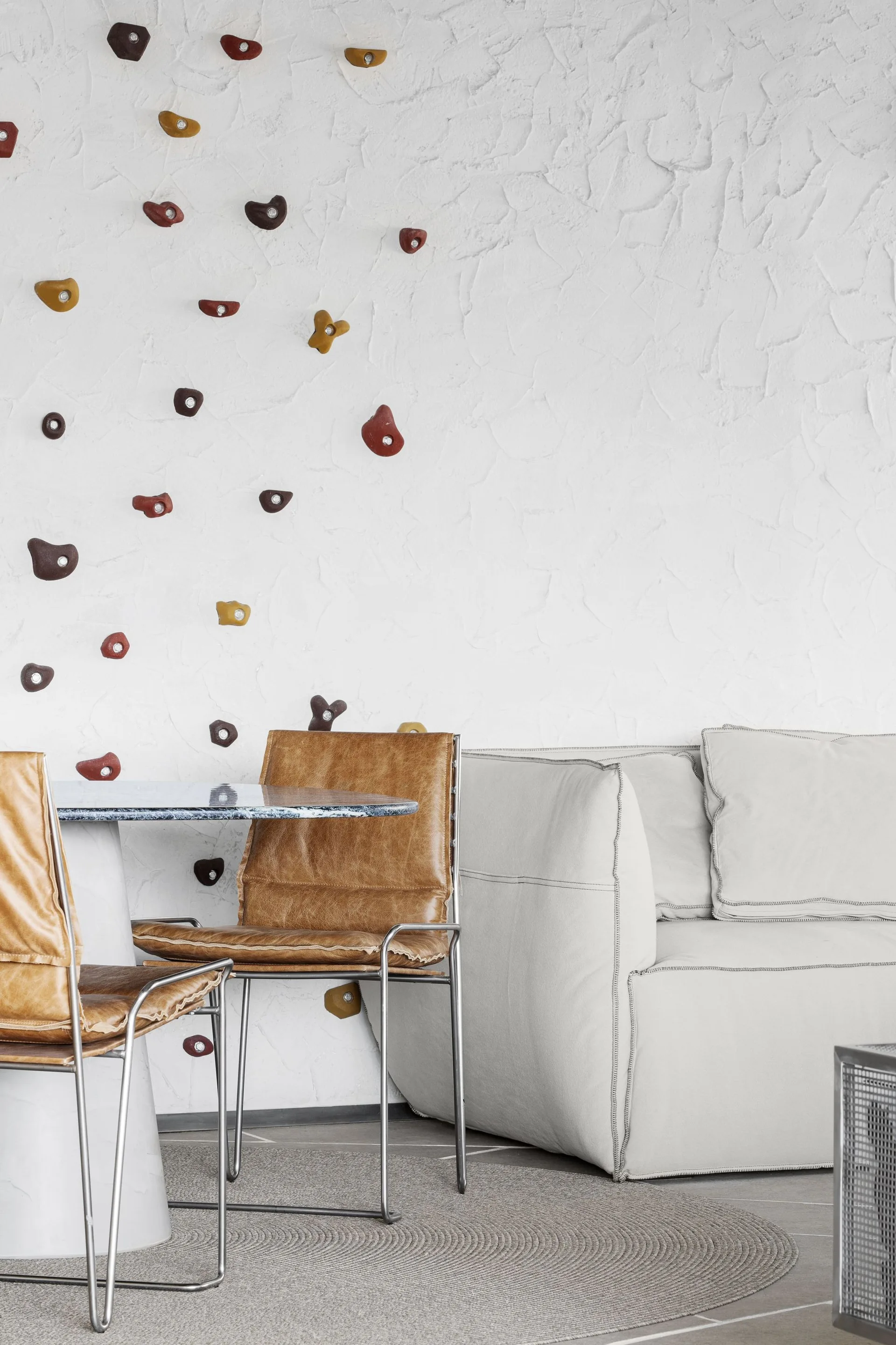 A modern living area with a white sofa and tan leather chairs at a round table set against a textured white wall with multi-colored climbing holds.