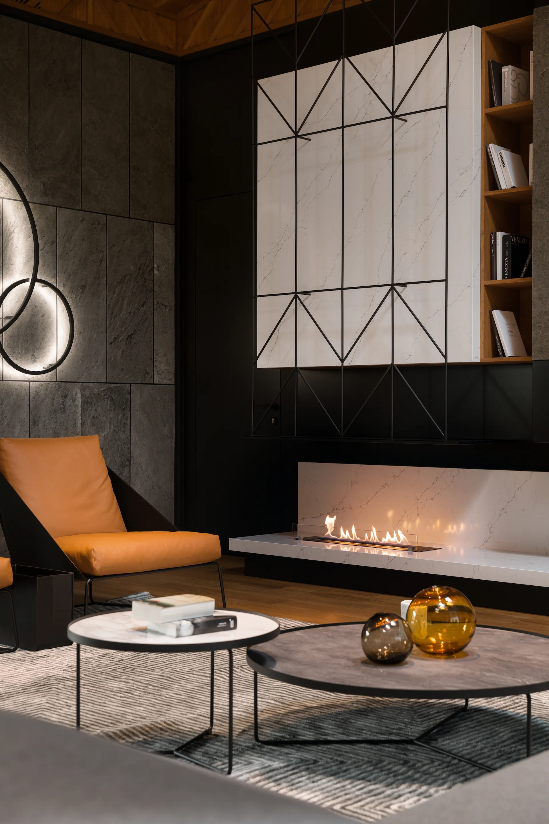 Modern living room with a linear fireplace on a white marble hearth, an orange leather chair, stone wall panels, and nesting coffee tables.
