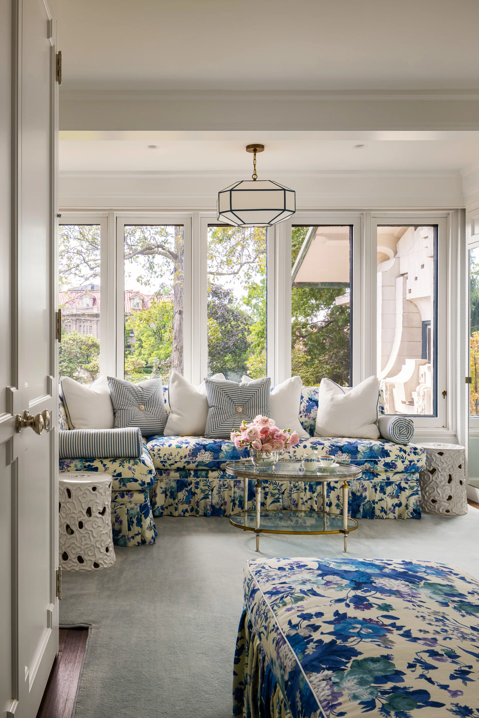 A bright sunroom features a blue and white floral upholstered sectional sofa flanked by white ceramic garden stools beneath a row of large windows.