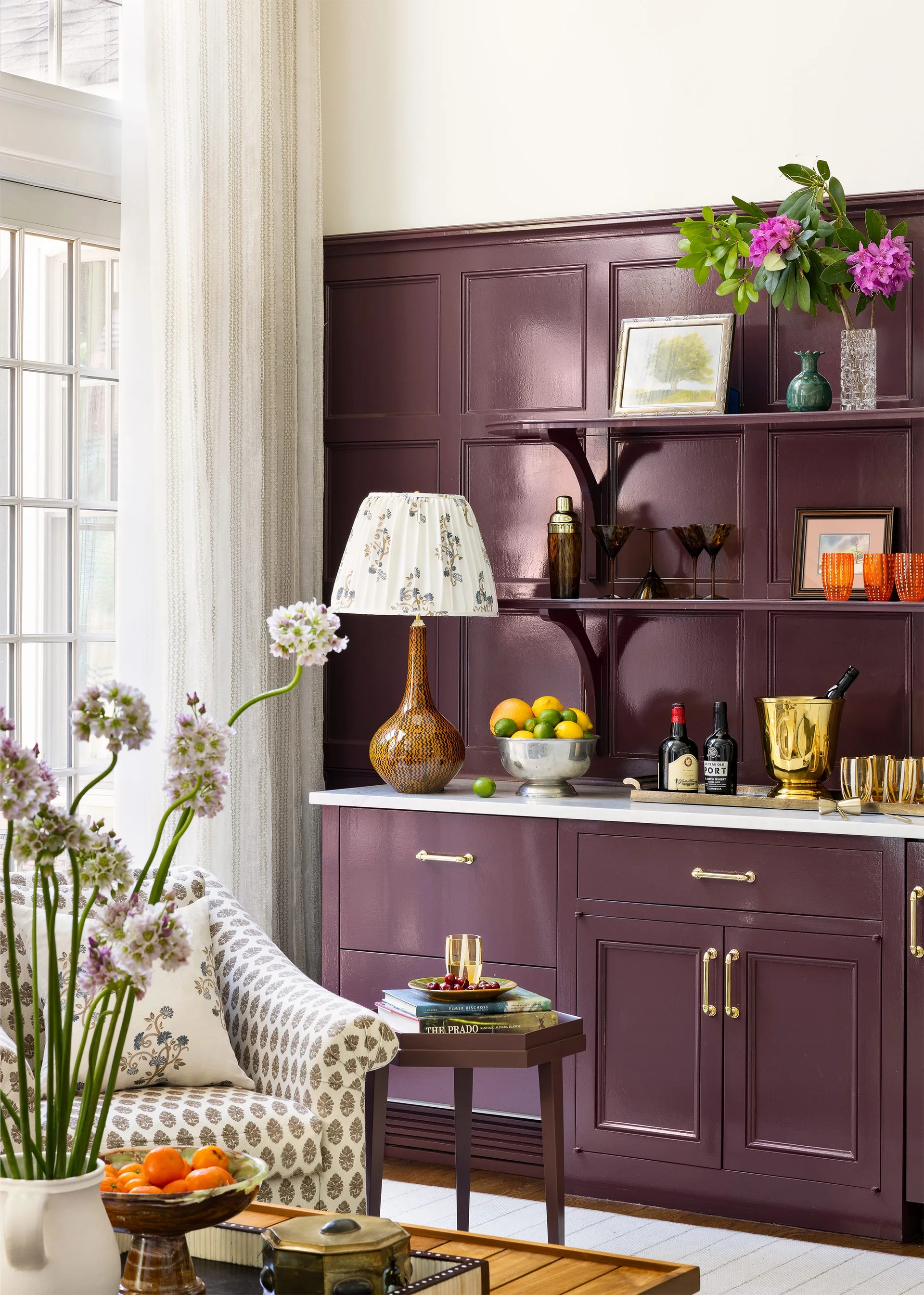 A glossy deep purple built-in bar cabinet features gold hardware, marble countertops, open shelves with glassware, and a patterned armchair nearby.