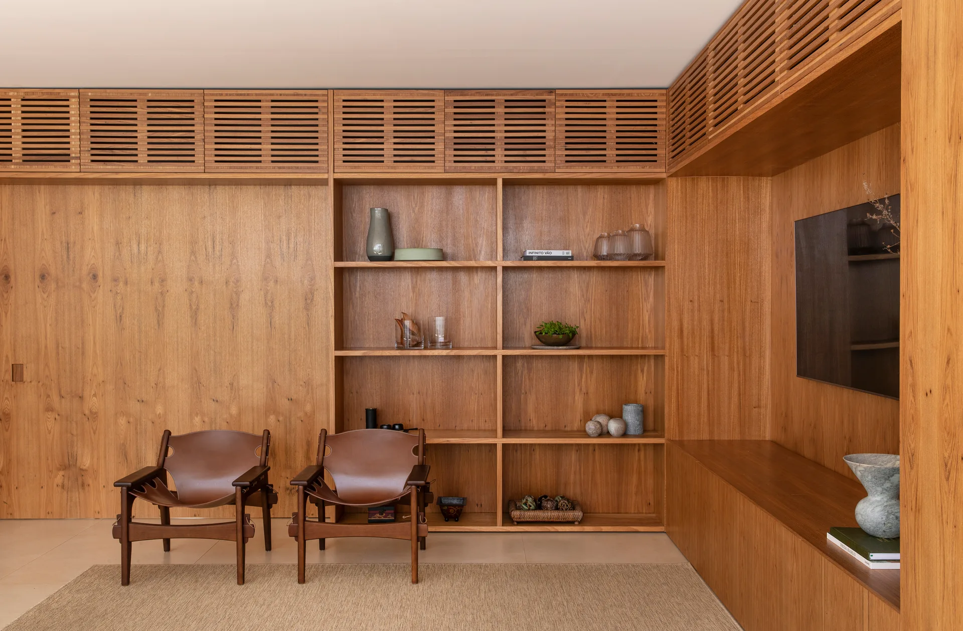 Two brown leather armchairs stand before a custom wood wall paneling system featuring built-in shelves, slatted cabinets, and an integrated TV.