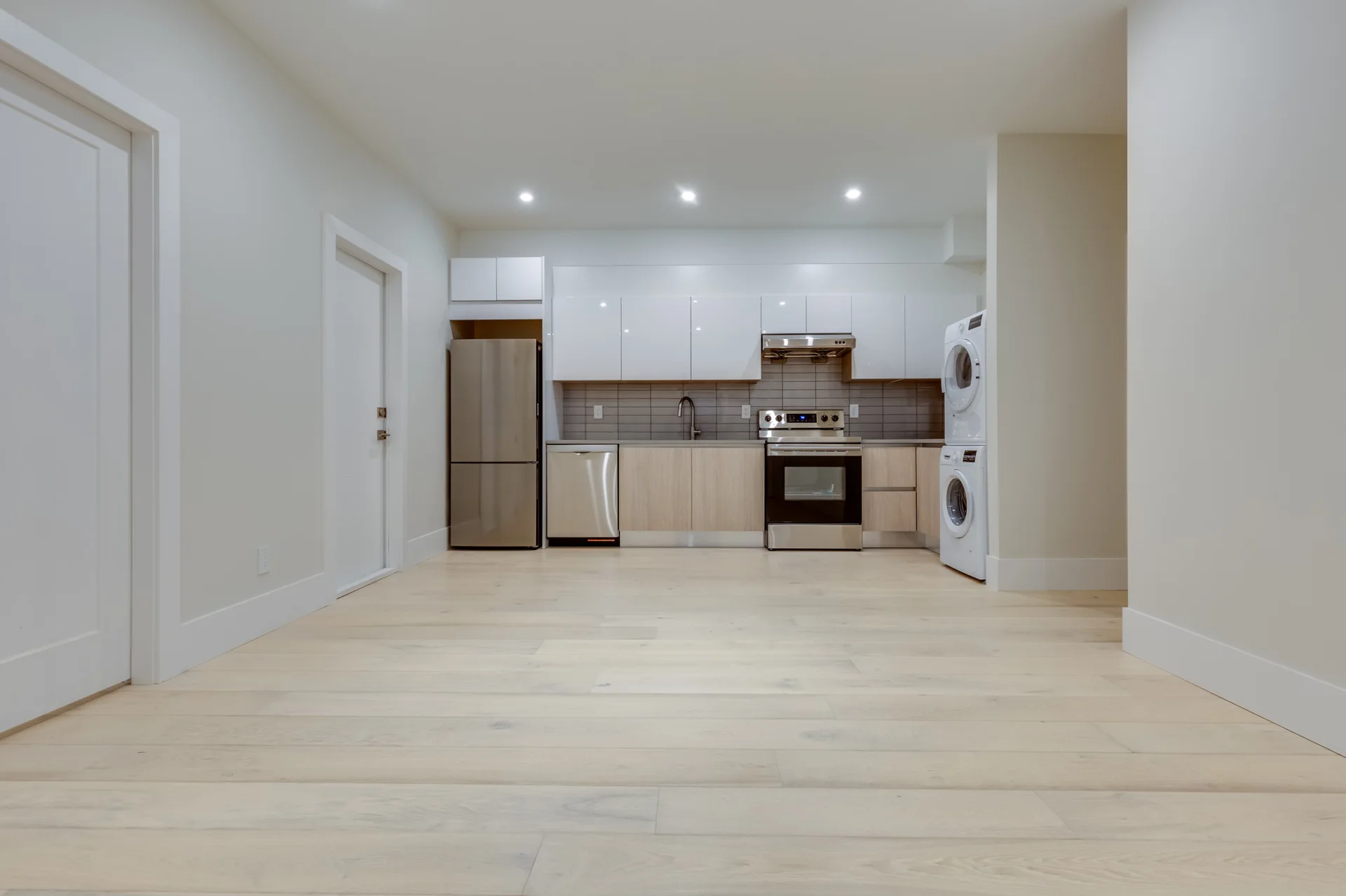 A modern kitchen features dual-tone wood and white cabinets, stainless steel appliances, a grey tile backsplash, and a stacked white washer and dryer.