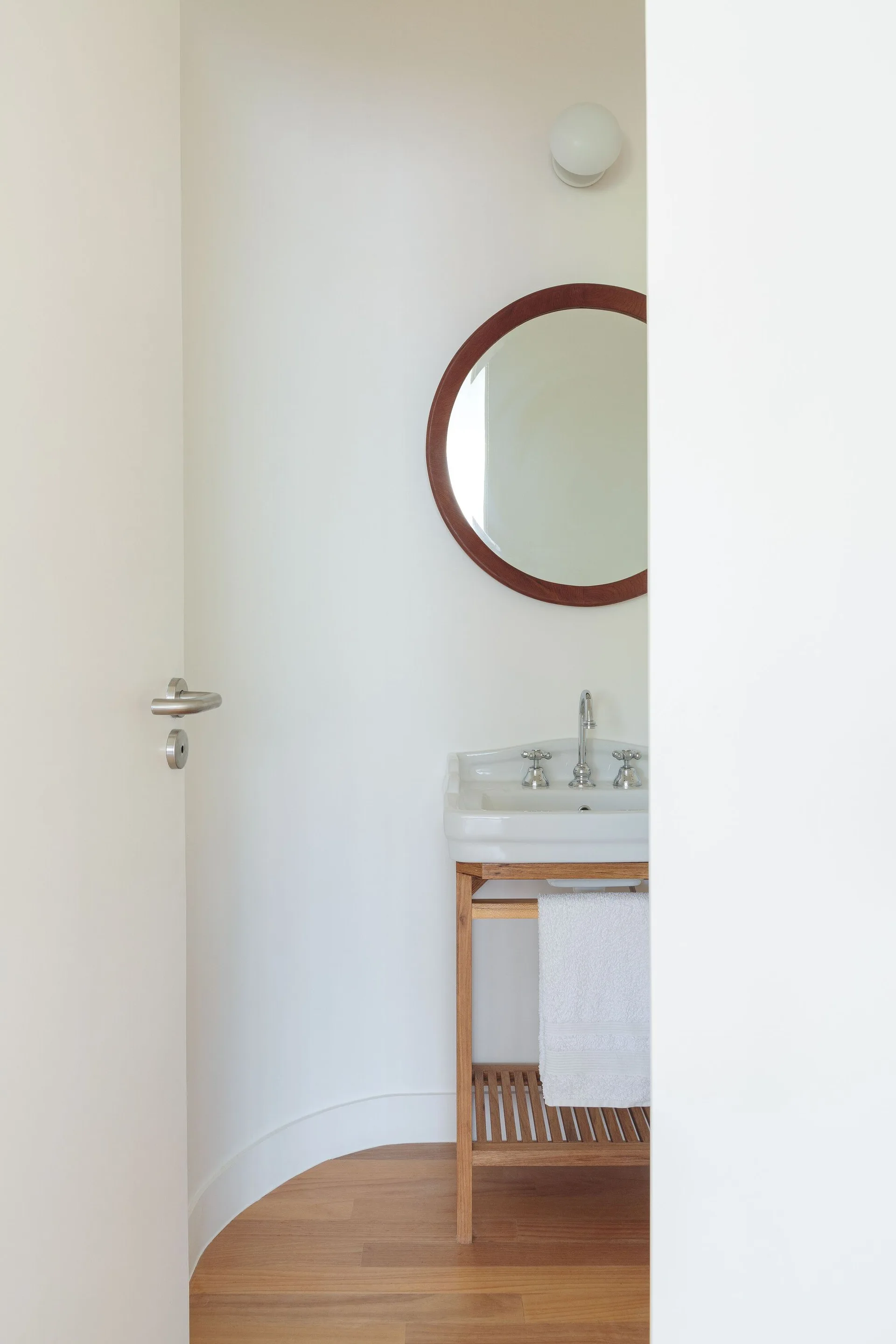 A small powder room features a white porcelain sink on a wooden stand, a round wood-framed mirror, and a wall-mounted glass globe light.