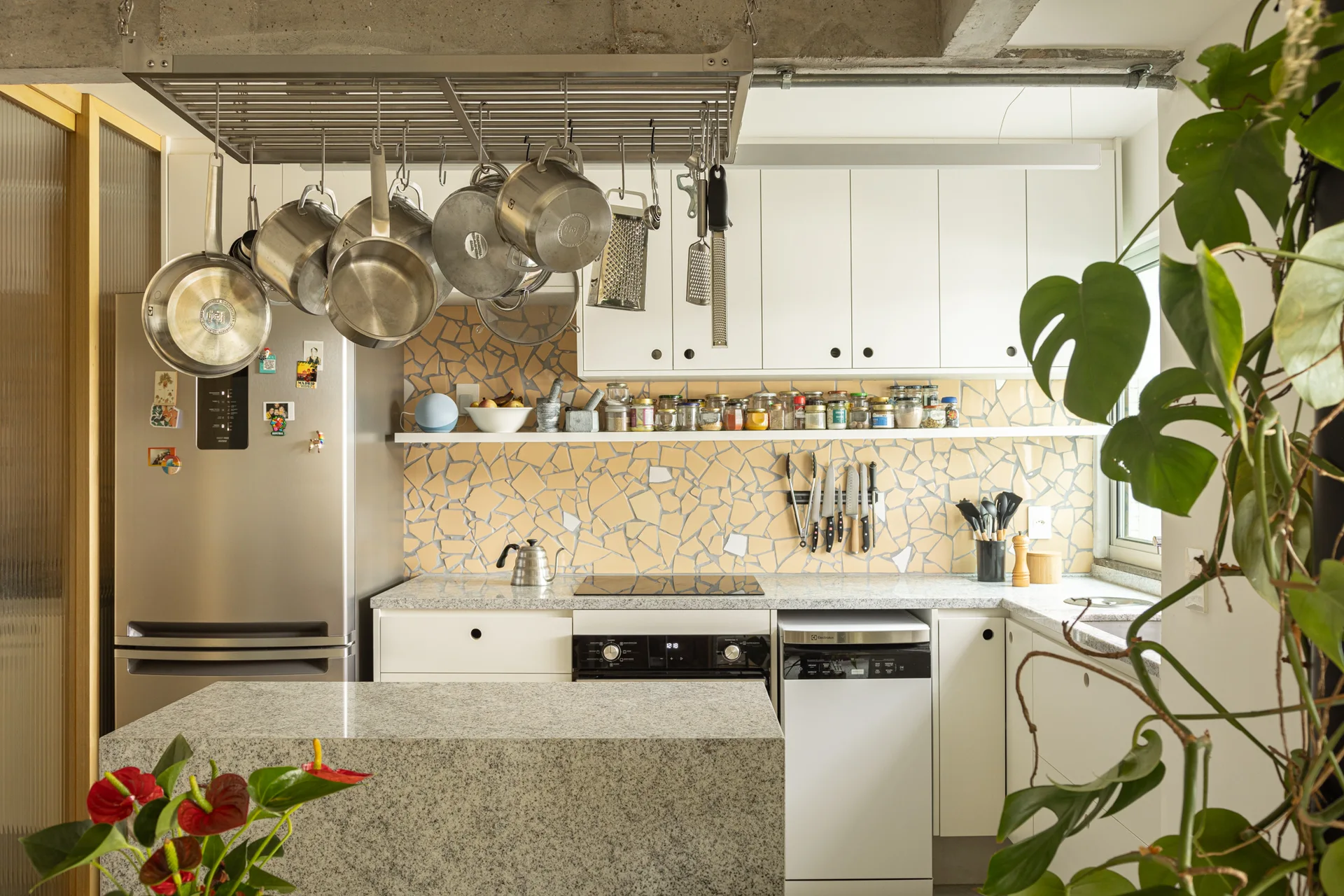 A kitchen features a yellow mosaic tile backsplash, white flat-front cabinetry, a granite island, and stainless steel pots hanging from a ceiling rack.