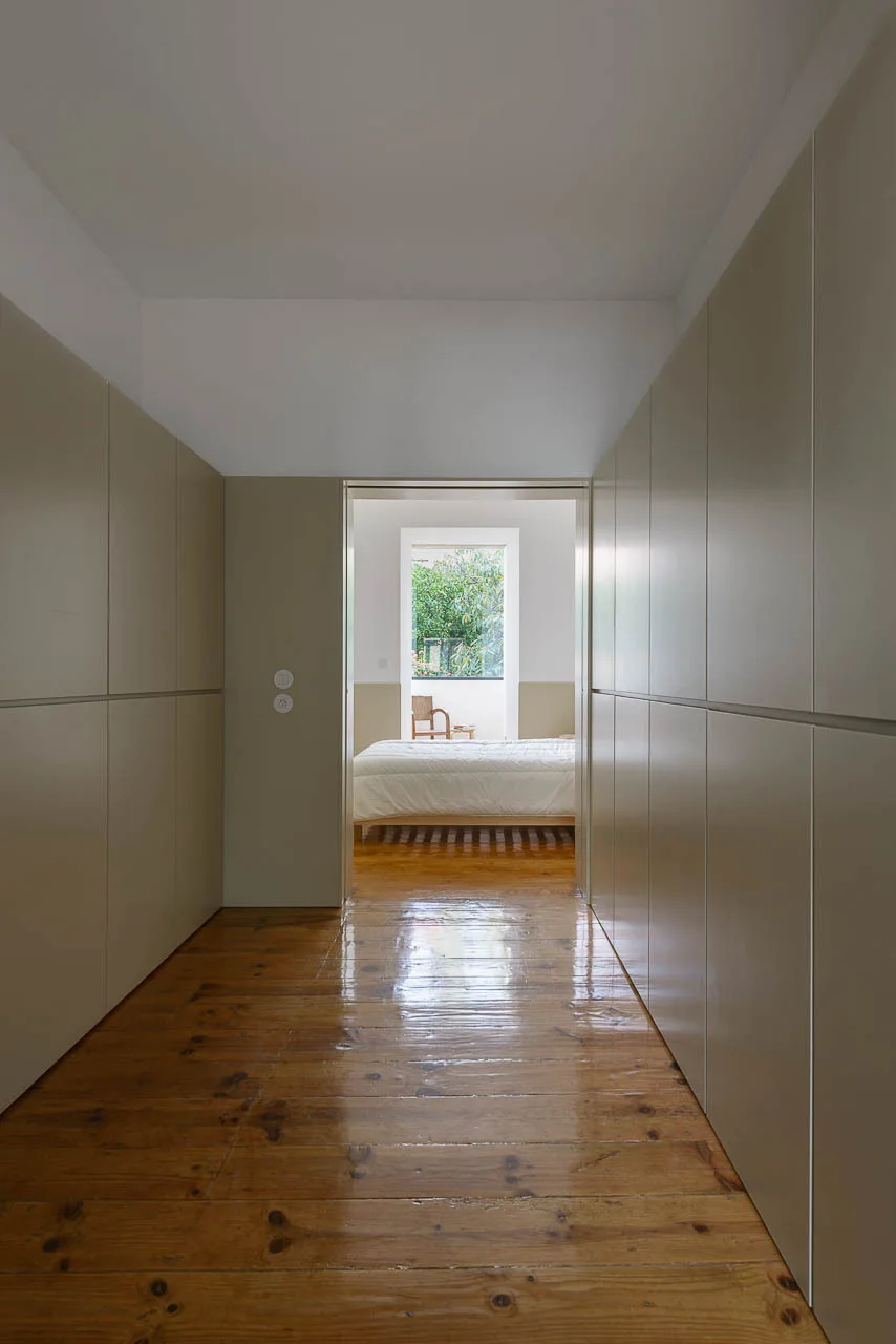A hallway flanked by tall, seamless beige wardrobes leads into a sunlit bedroom with glossy wood plank flooring.