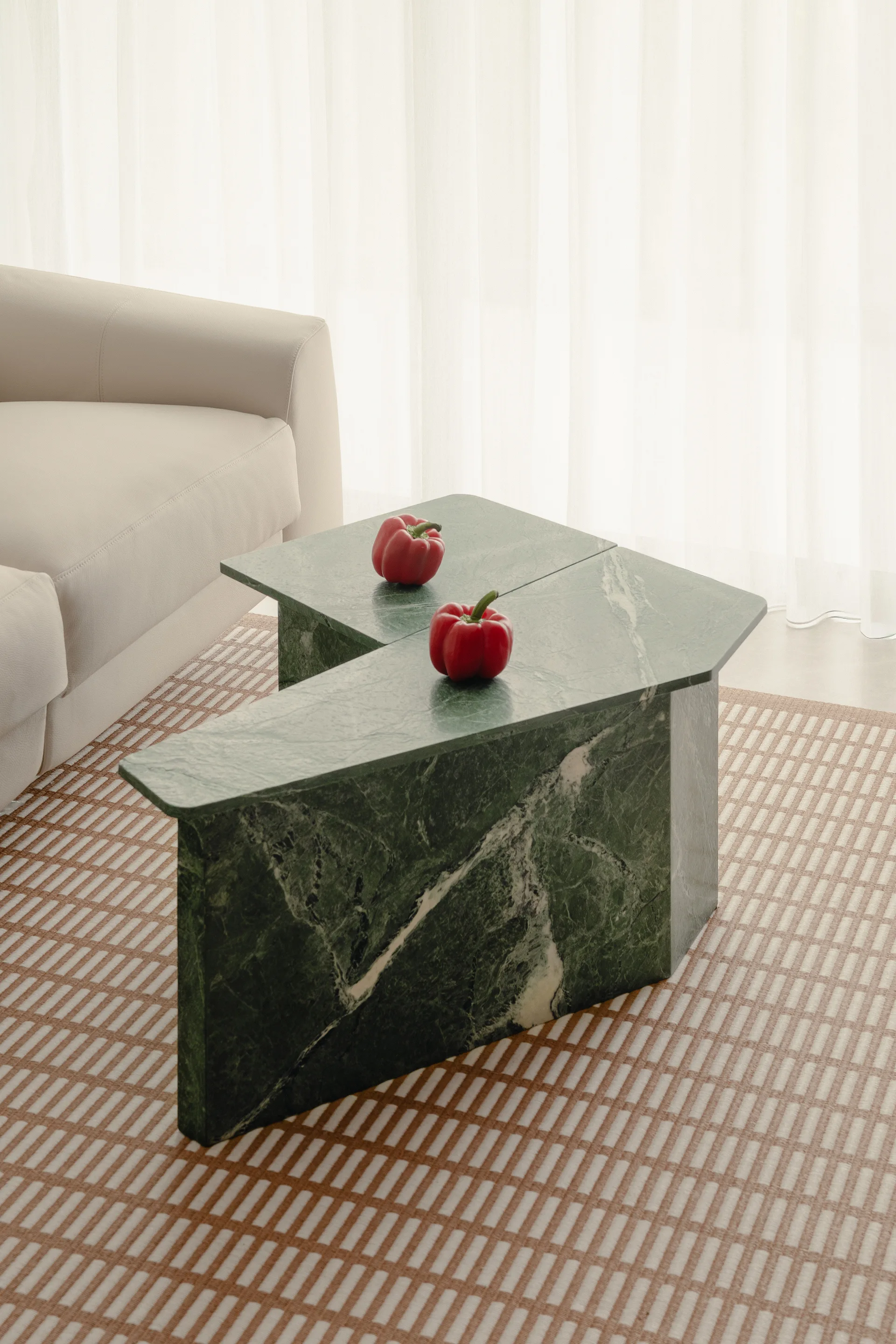 A geometric green marble coffee table topped with two red bell peppers sits on a grid-patterned rug next to a beige sofa.