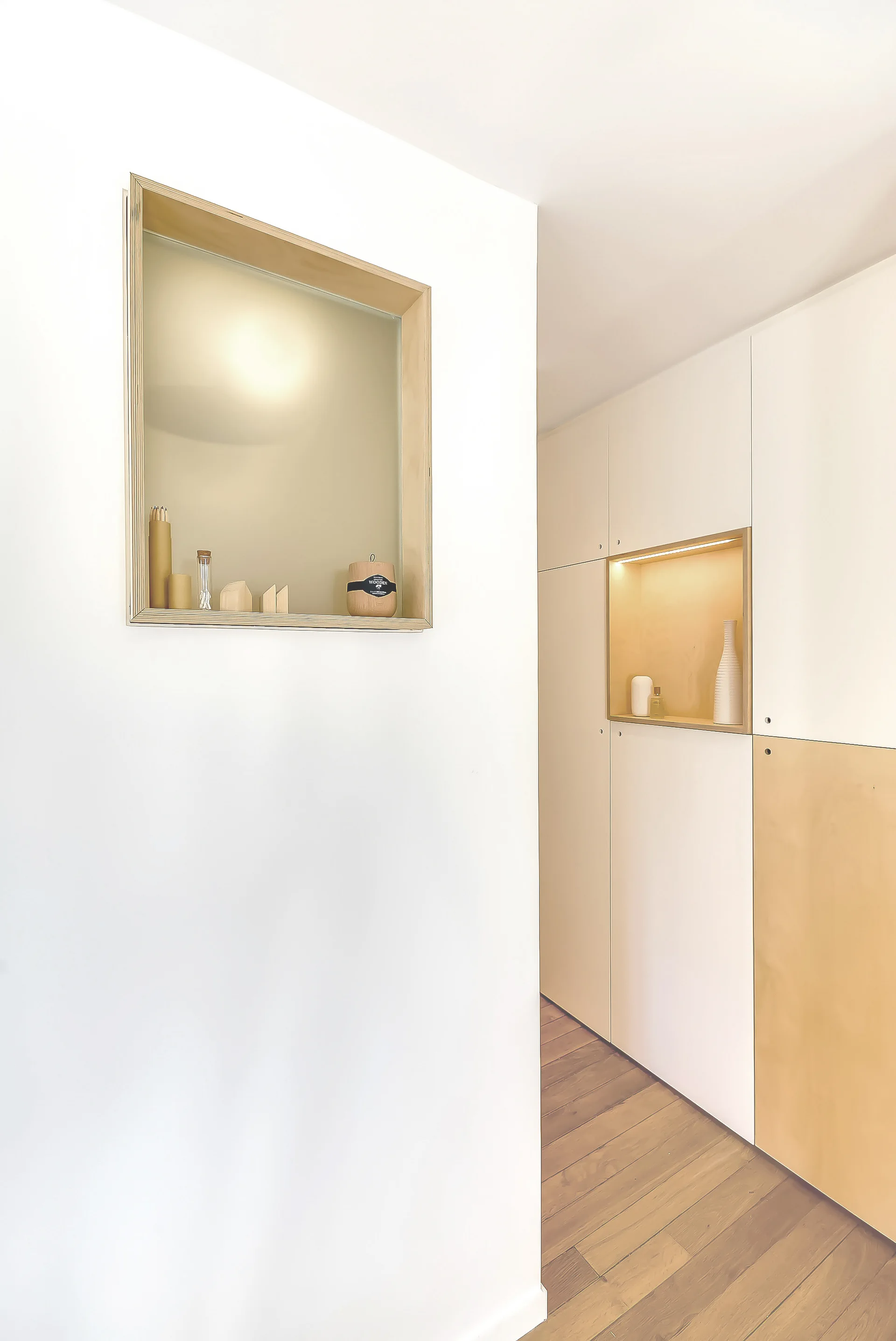 A minimalist white hallway featuring custom built-in cabinetry with warm wood niches, integrated lighting, and medium-brown wood plank flooring.