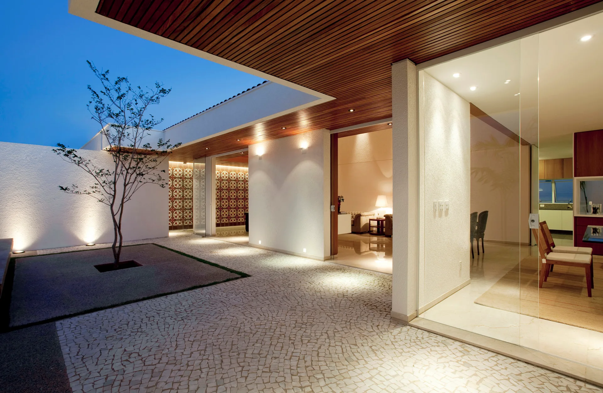A modern outdoor patio featuring cobblestone paving, stucco walls, and a slatted wood ceiling that looks into warmly lit interiors through glass walls.