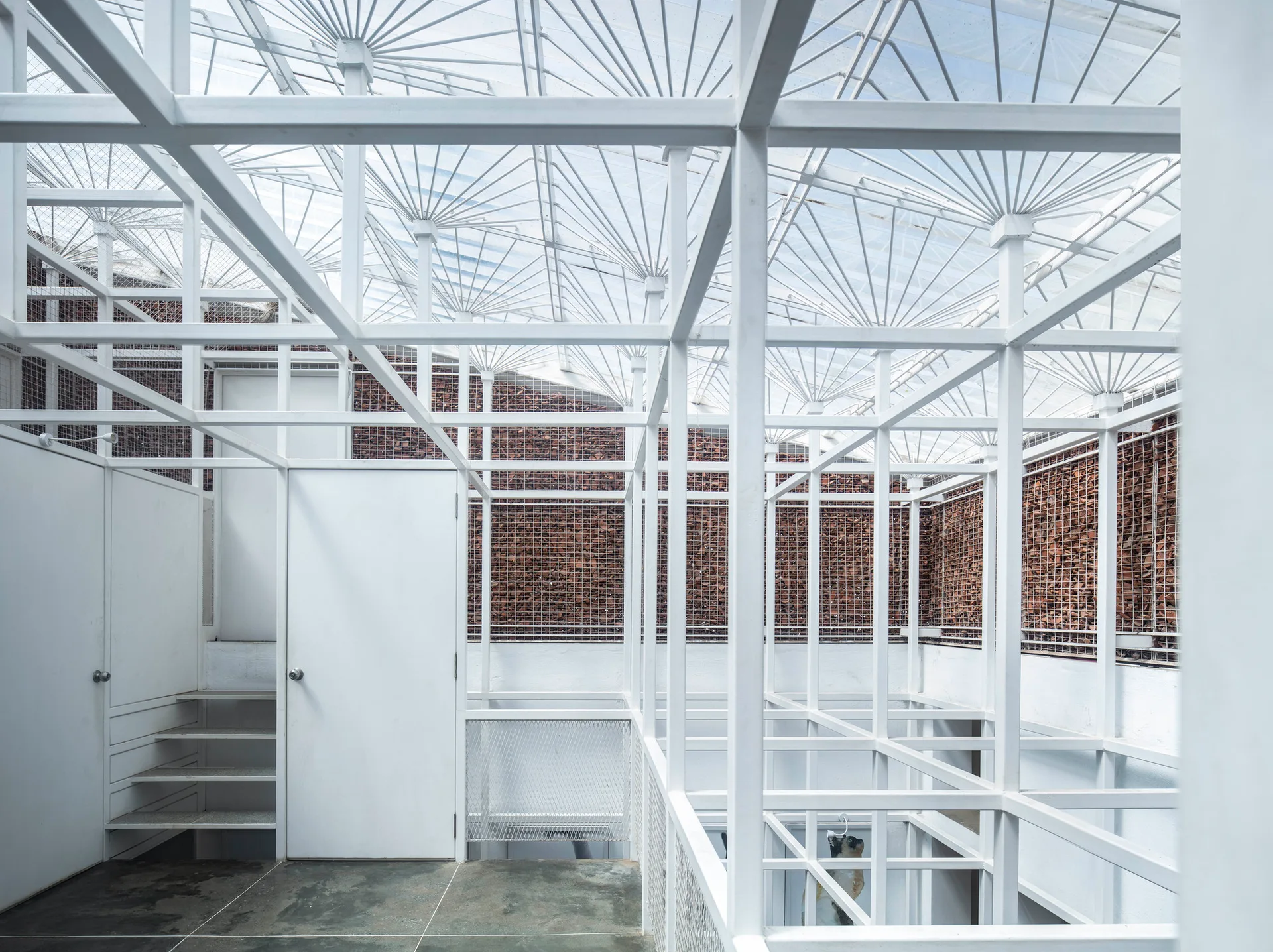 An interior space defined by a white metal grid structure, featuring fan-patterned ceiling supports and gabion stone walls.