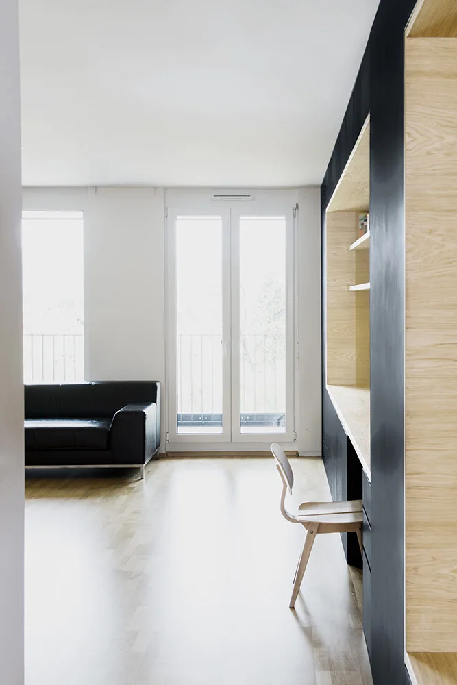 A minimalist space with light wood floors features a matte black wall unit with a built-in desk, a wooden chair, and a black leather sofa.