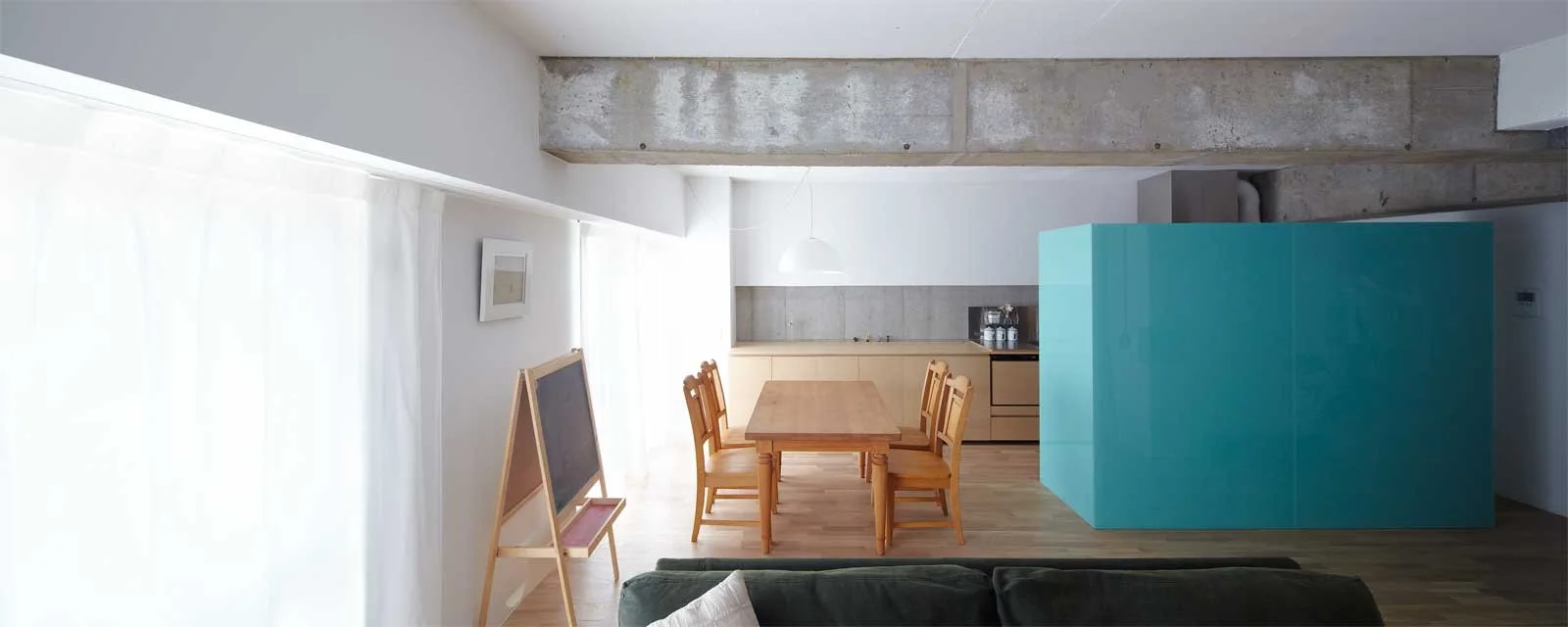 An open dining and kitchen area featuring a wooden table, a raw concrete ceiling beam, and a large glossy turquoise room divider.